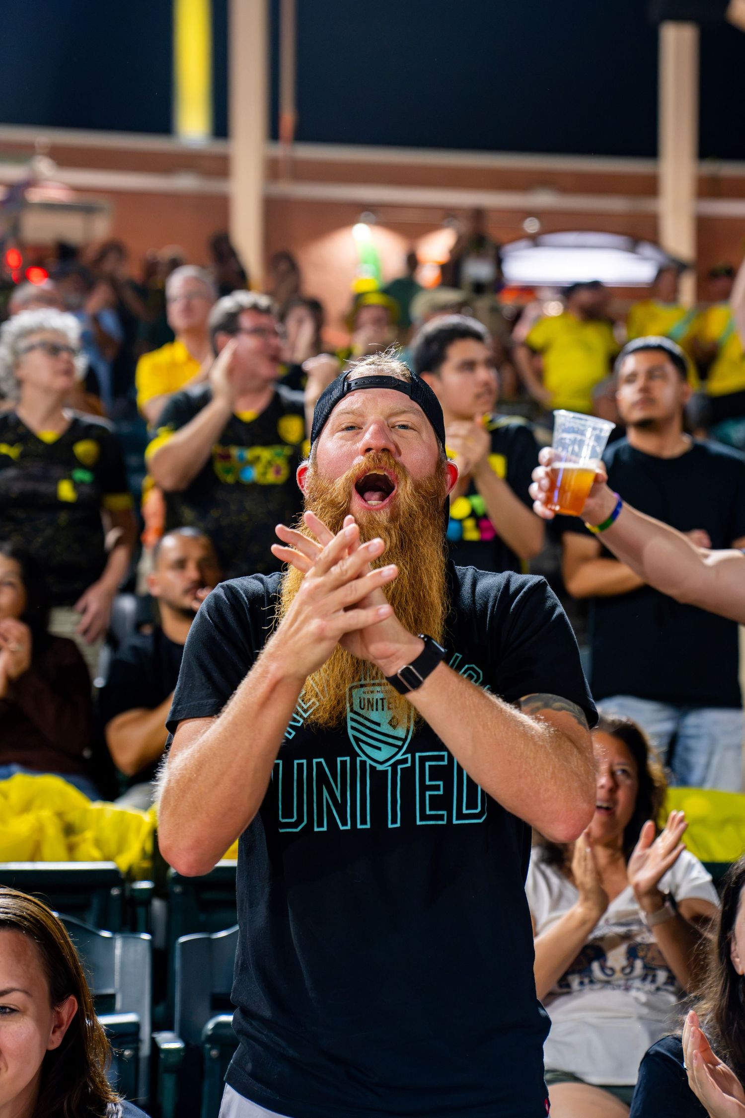 Enthusiastic bearded male soccer fan in a United shirt clapping and cheering at a crowded stadium event.