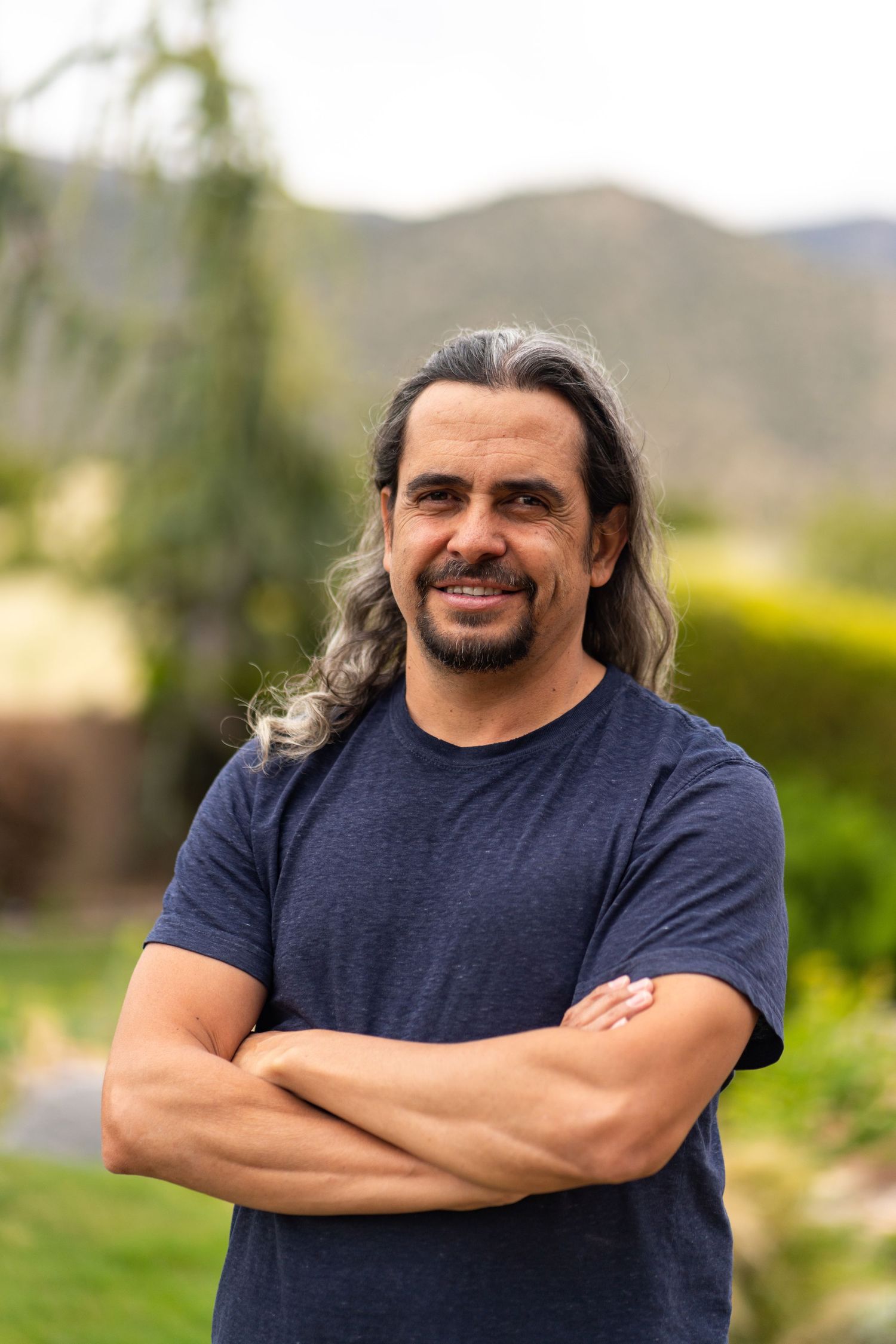 Professional headshot of a person in a navy t-shirt with long hair outdoors against a mountain backdrop.