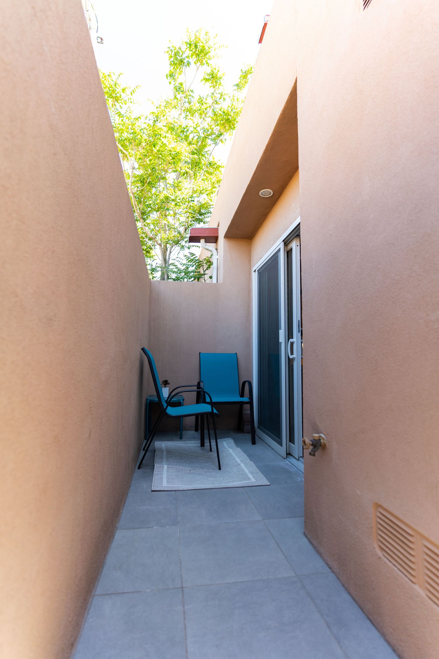 Outdoor patio corridor with tan stucco walls, concrete flooring, and black chairs visible at the end.