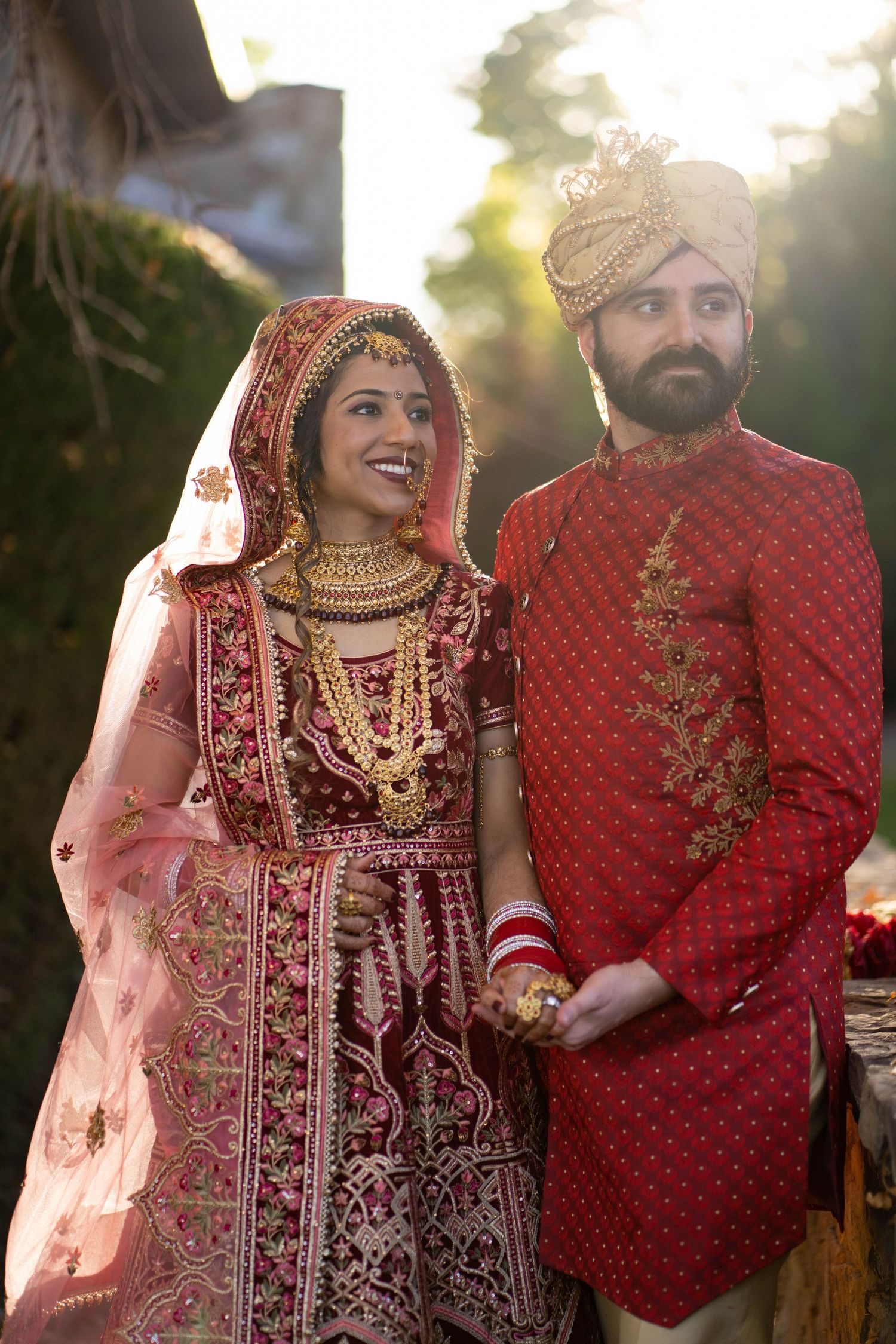 A couple in ornate red wedding attire poses together during golden hour, with detailed embroidery on their traditional clothes.