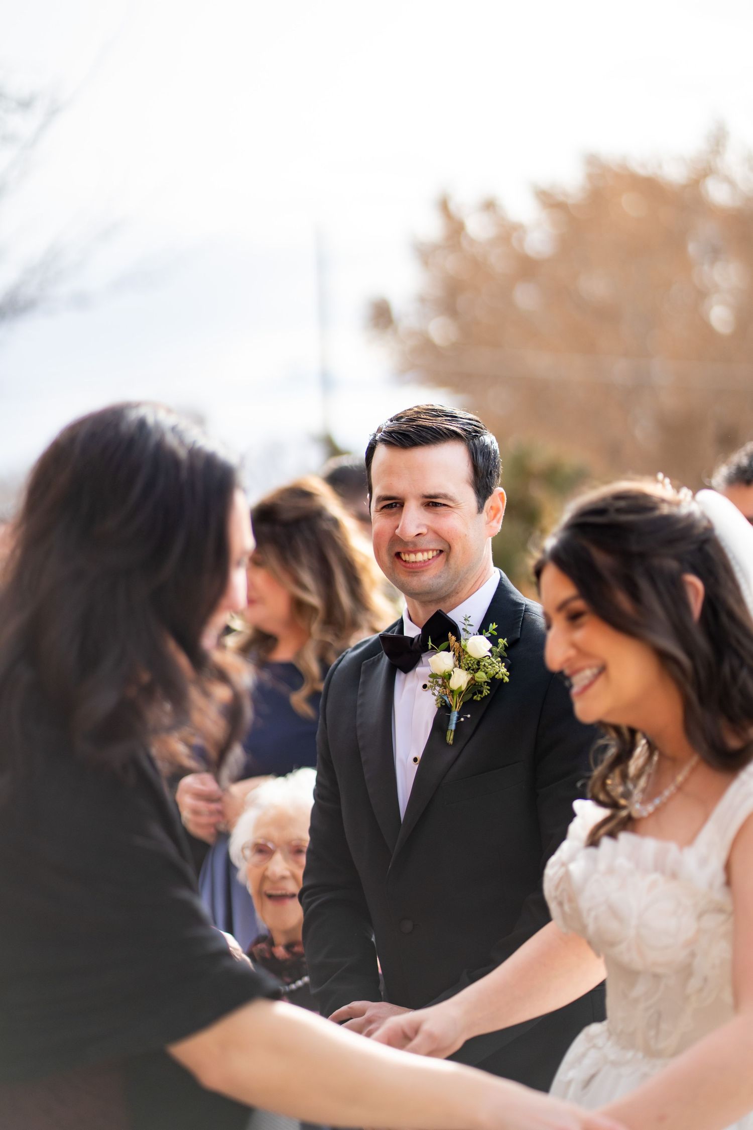 Smiling groom in black tuxedo with floral boutonniere stands beside bride during outdoor wedding ceremony.