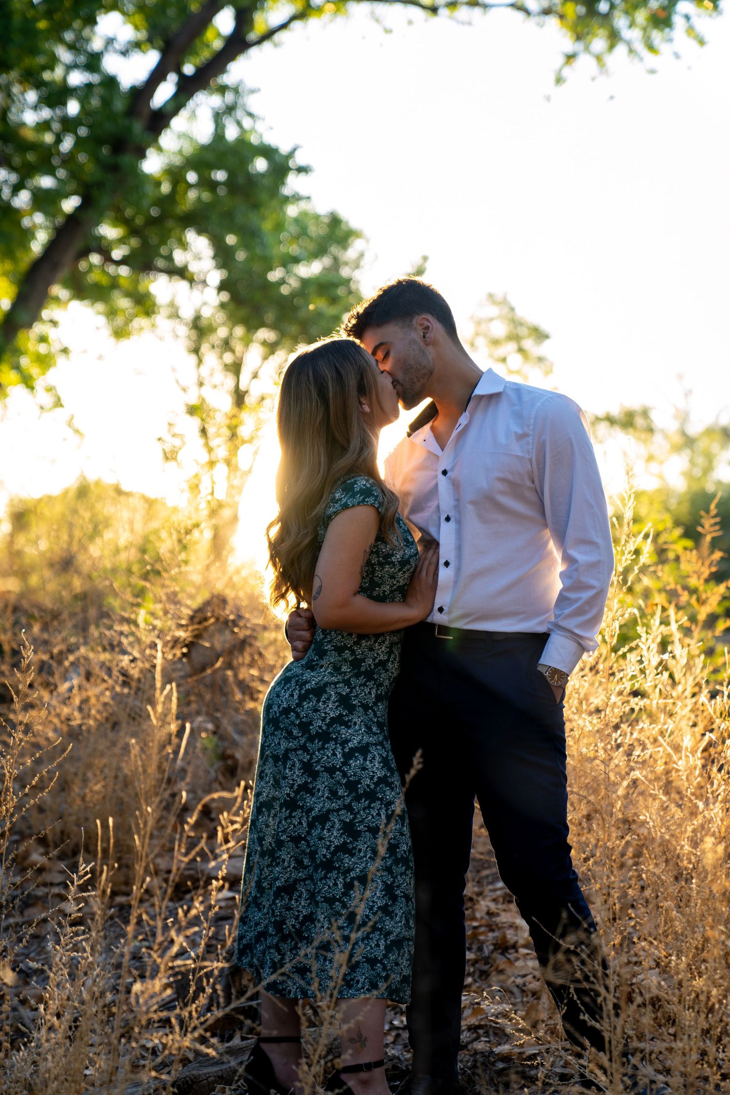 A romantic couple embraces in golden sunlight among tall dried grass during a sunset photoshoot.