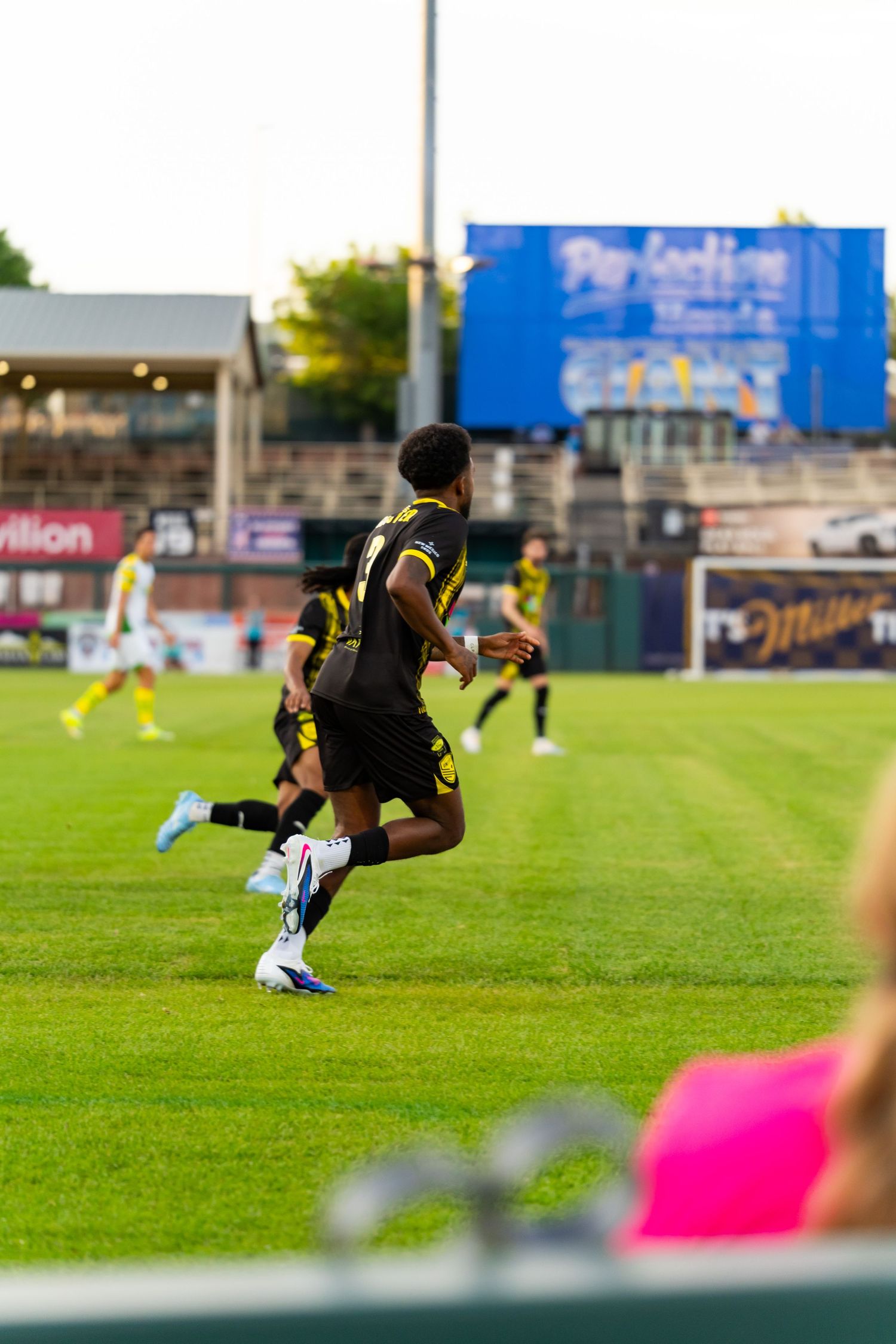 Soccer players in black and yellow uniforms run on a green field during an evening match at a stadium.