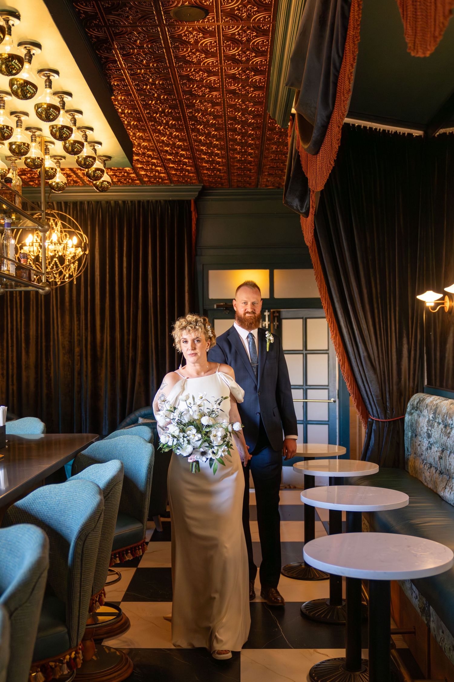 Bride in white gown holding bouquet walks with groom in navy suit through elegant restaurant venue.