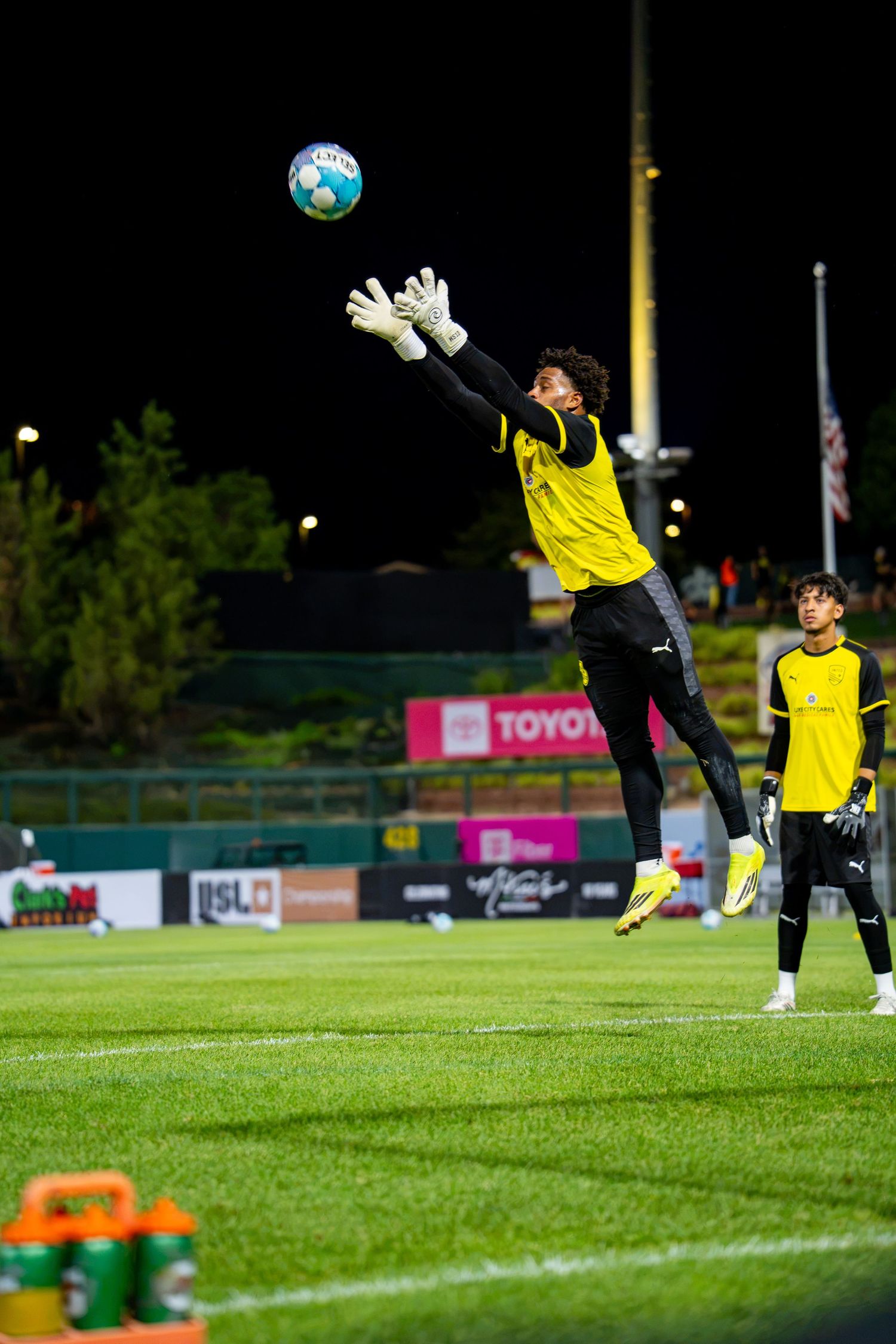 Goalkeeper in yellow jersey leaps to catch a soccer ball during a night match on a green field.