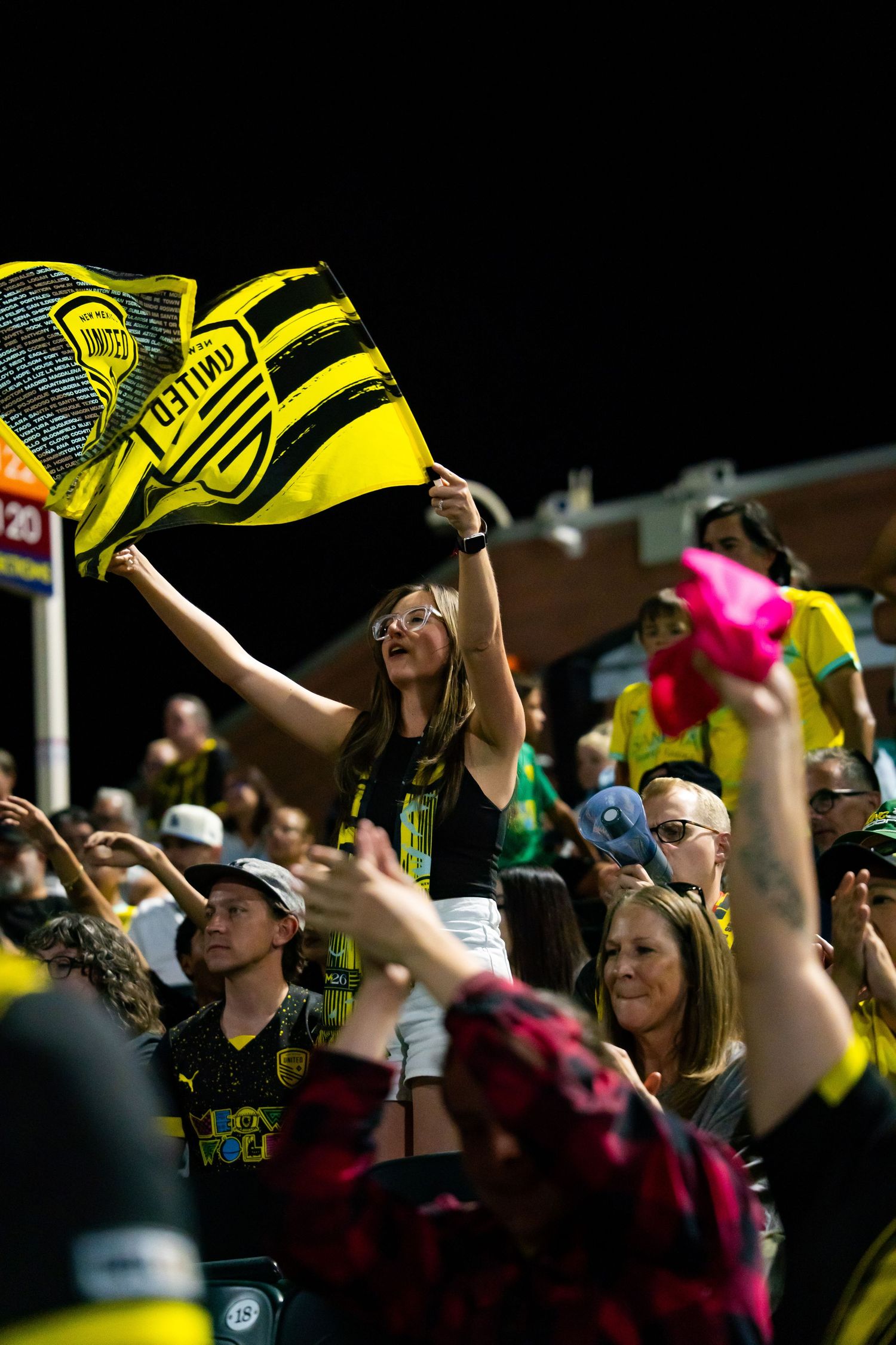 New Mexico United soccer fan waves yellow and black team flag in crowded stadium at night game.