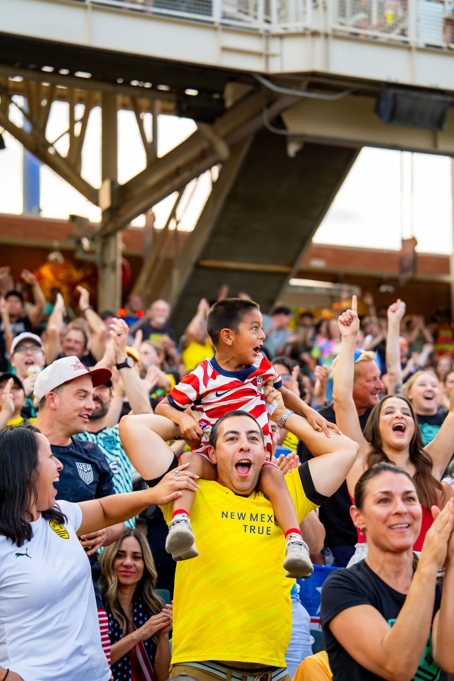 Excited soccer fans celebrate in stadium, man in yellow New Mexico True shirt lifts child on shoulders.