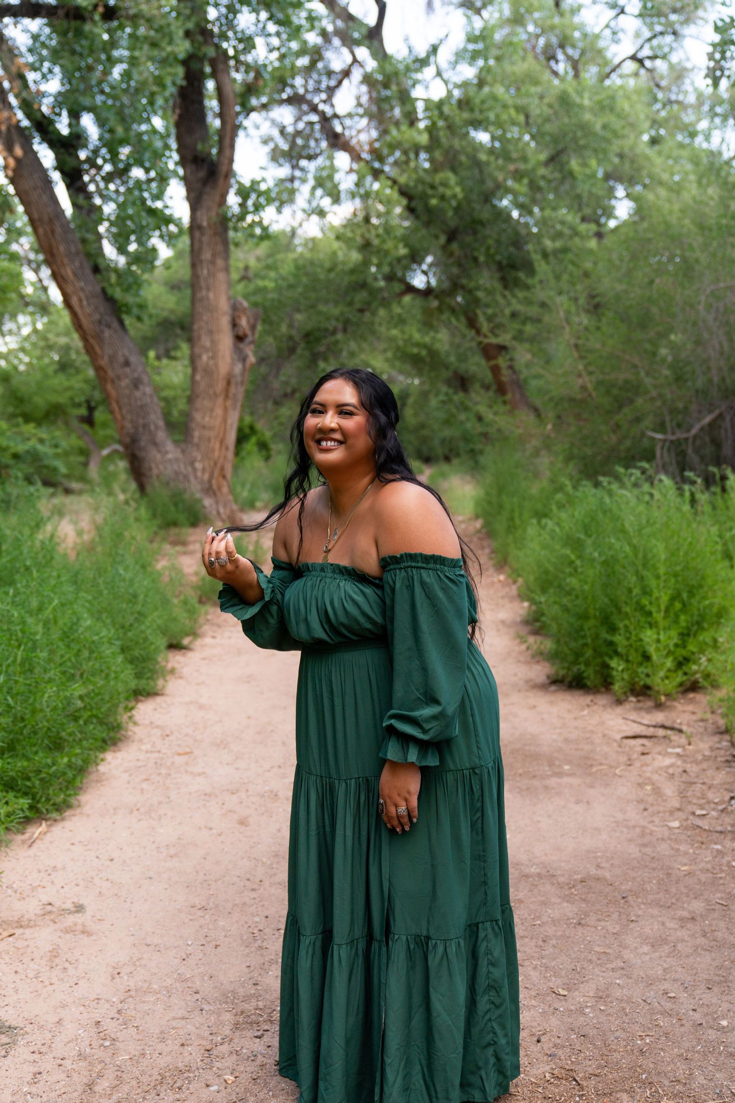 Woman in emerald green off-shoulder dress smiling on nature path.