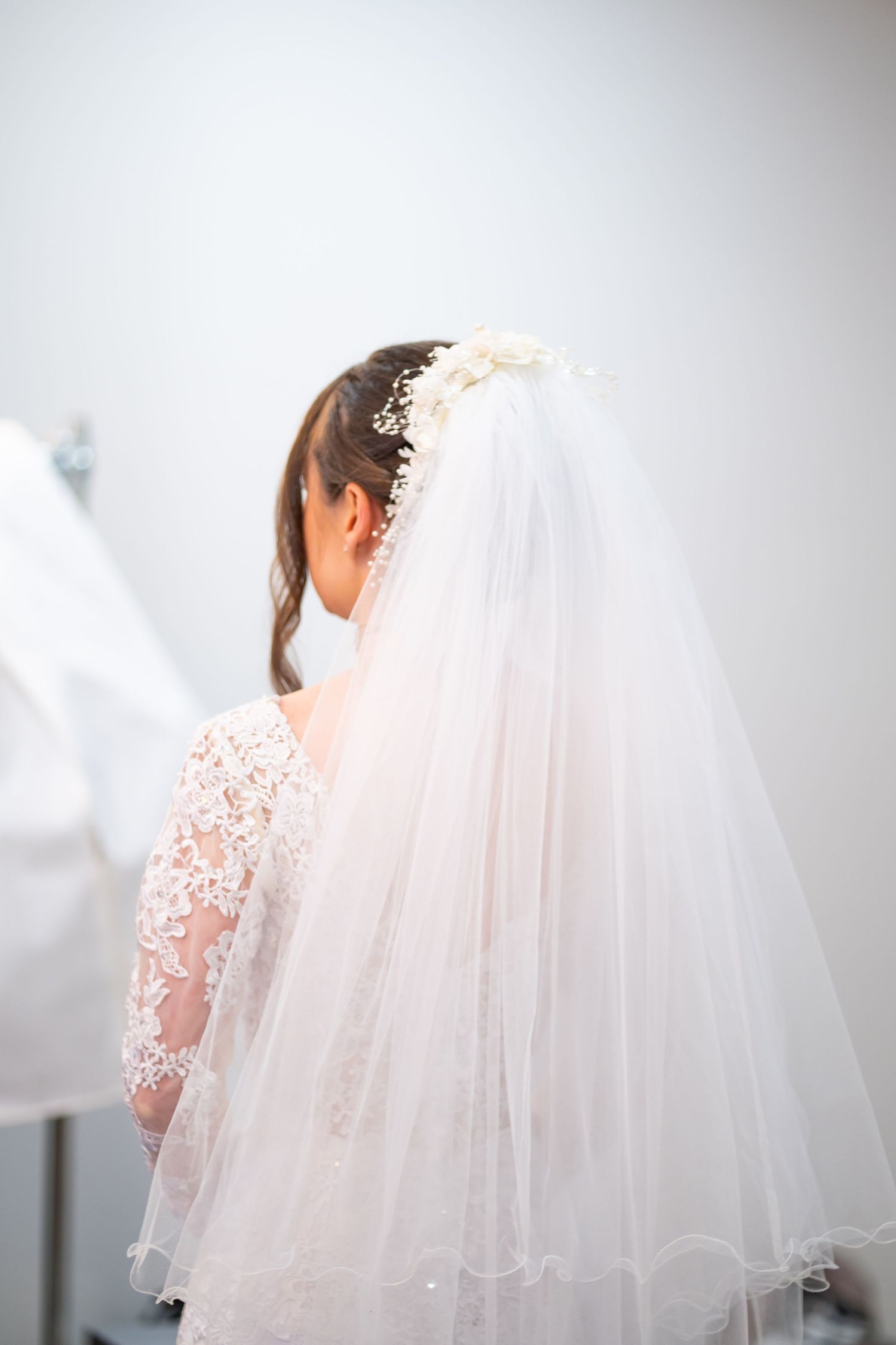 Bride in lace wedding dress with floral veil viewed from behind in bridal boutique.