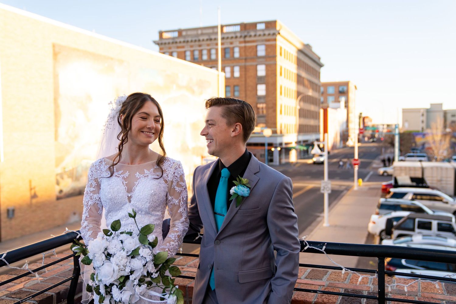 Bride in white lace gown and groom in gray suit share a romantic moment on a rooftop with city buildings at sunset.