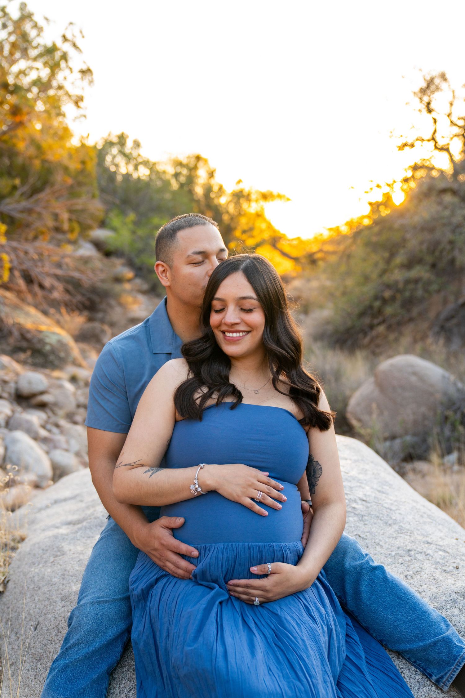 Couple in matching blue outfits sharing a tender maternity moment on rocks at golden hour sunset.