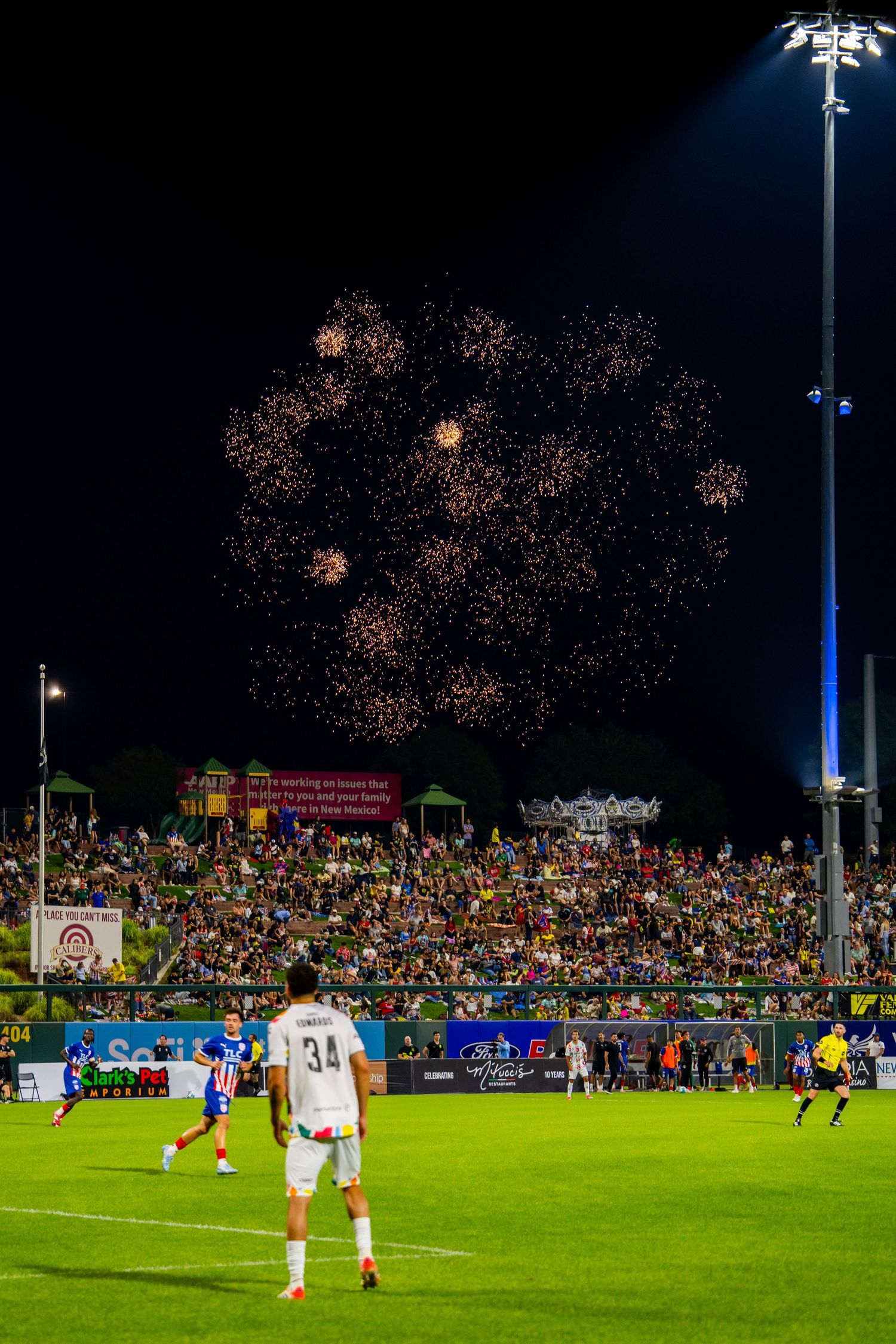 Soccer players on field with fireworks display lighting up night sky above packed stadium stands.