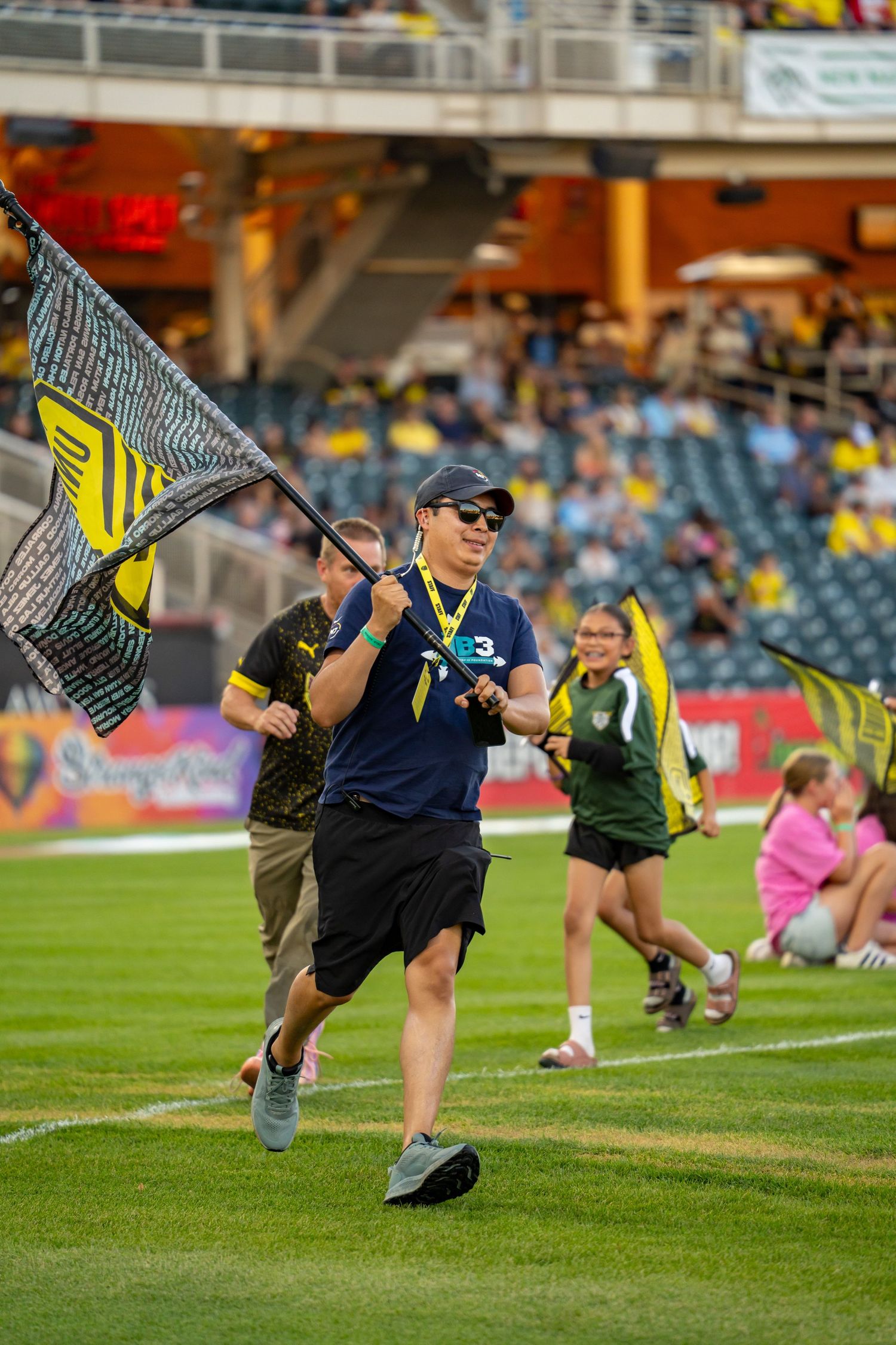 Fan runs across soccer field waving large black and yellow team flag during MLS match at packed stadium.