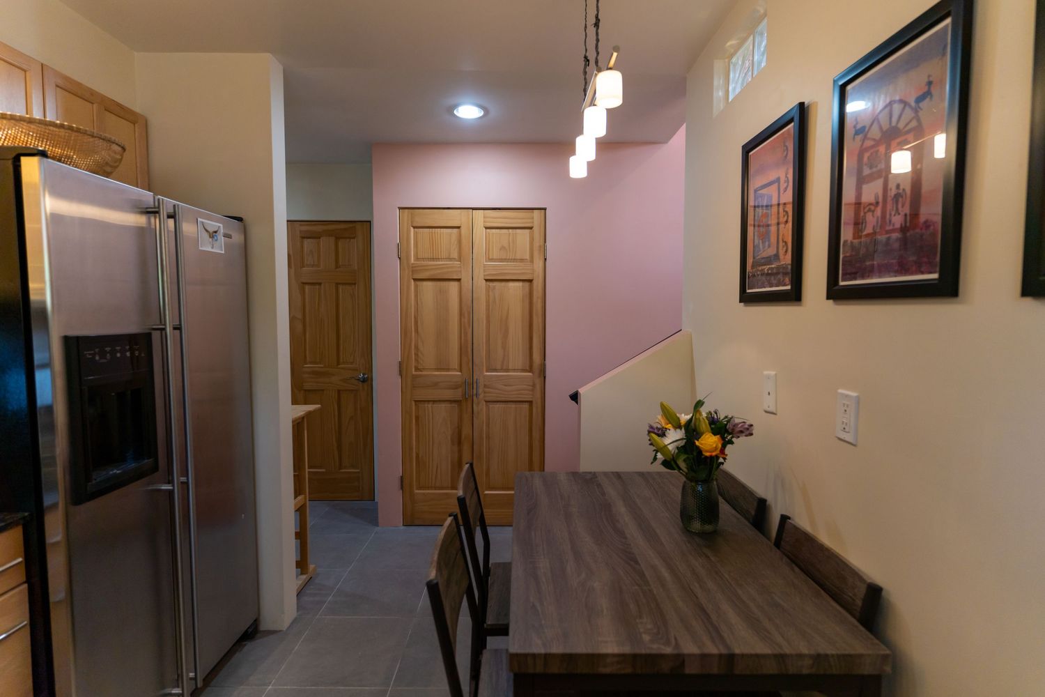 Modern kitchen dining area with stainless steel fridge, wooden doors, and pendant lighting over a gray table.