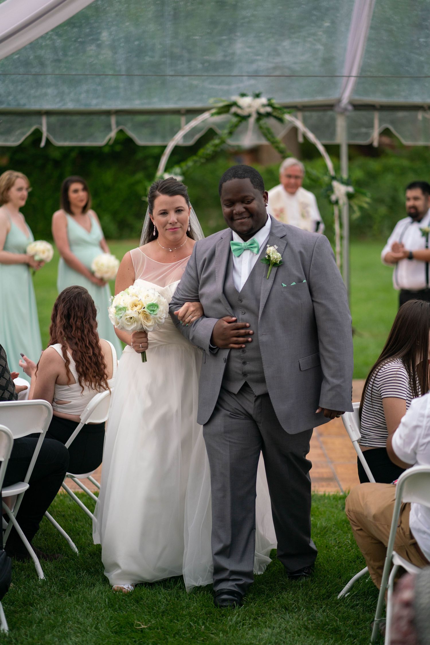 Wedding celebration under a white tent with guests seated on chairs on a grassy lawn.