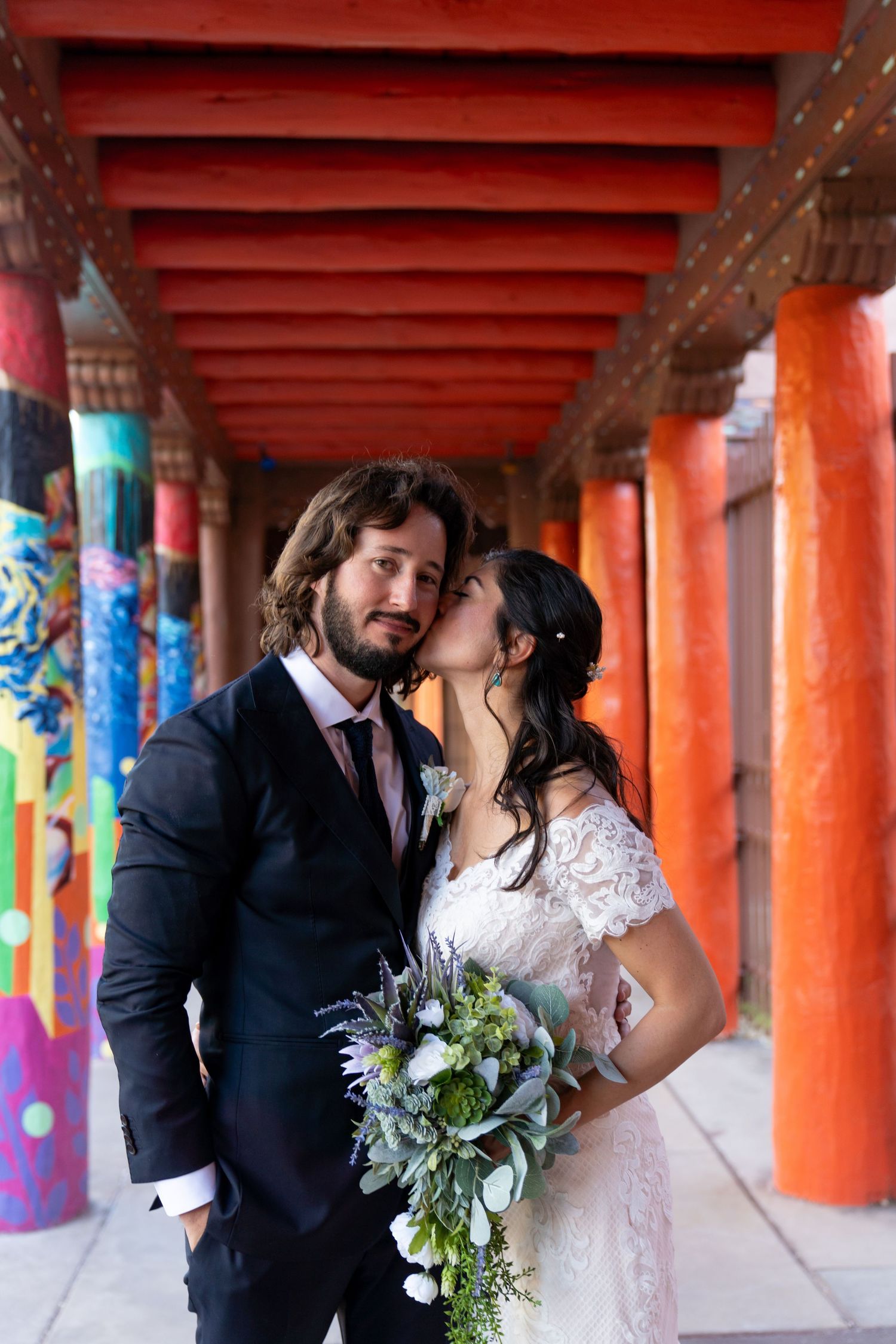 Wedding couple embraces under vibrant orange pillars with colorful murals.