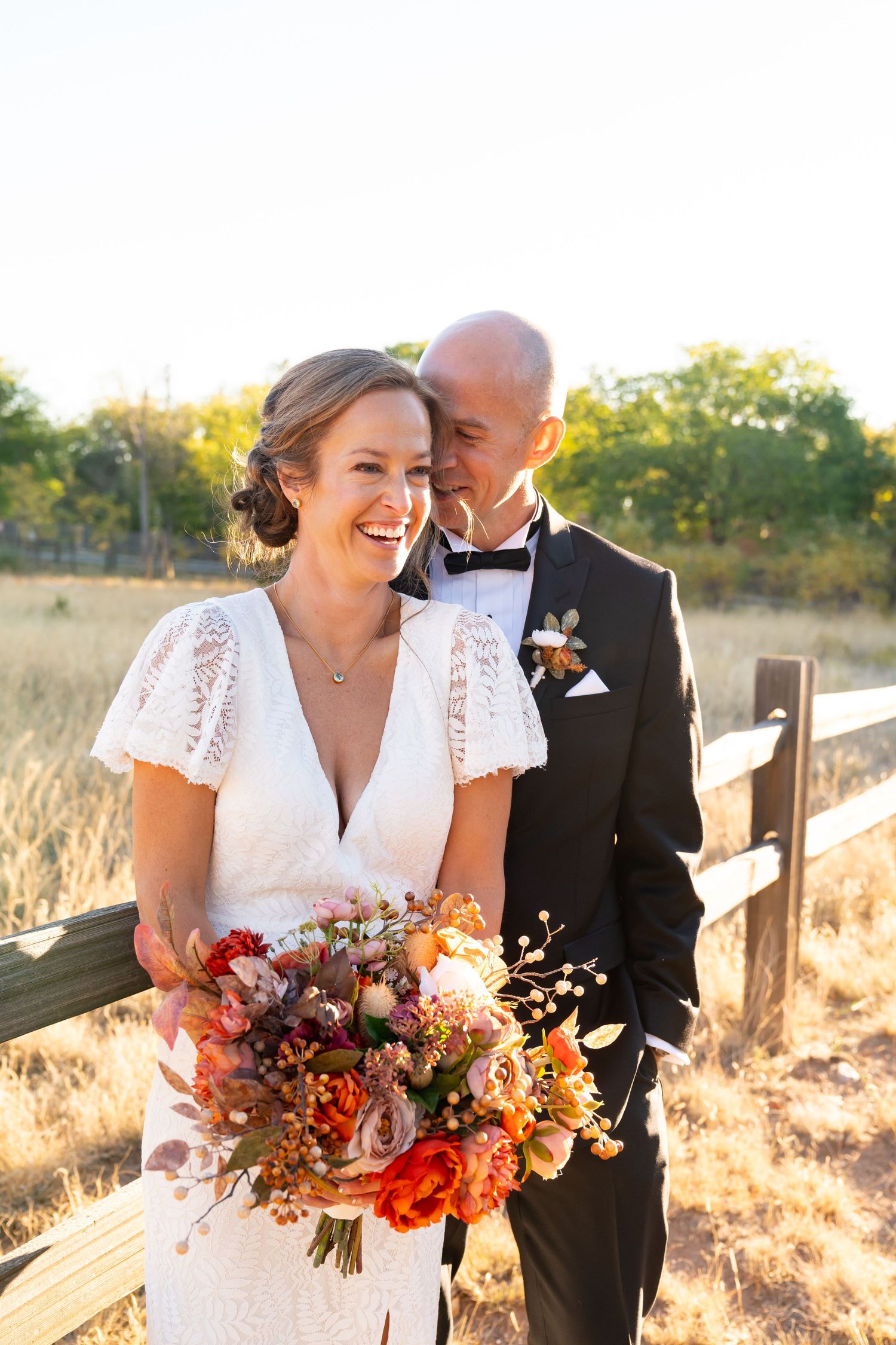 Newlyweds laugh together while holding vibrant fall bouquet near wooden fence.