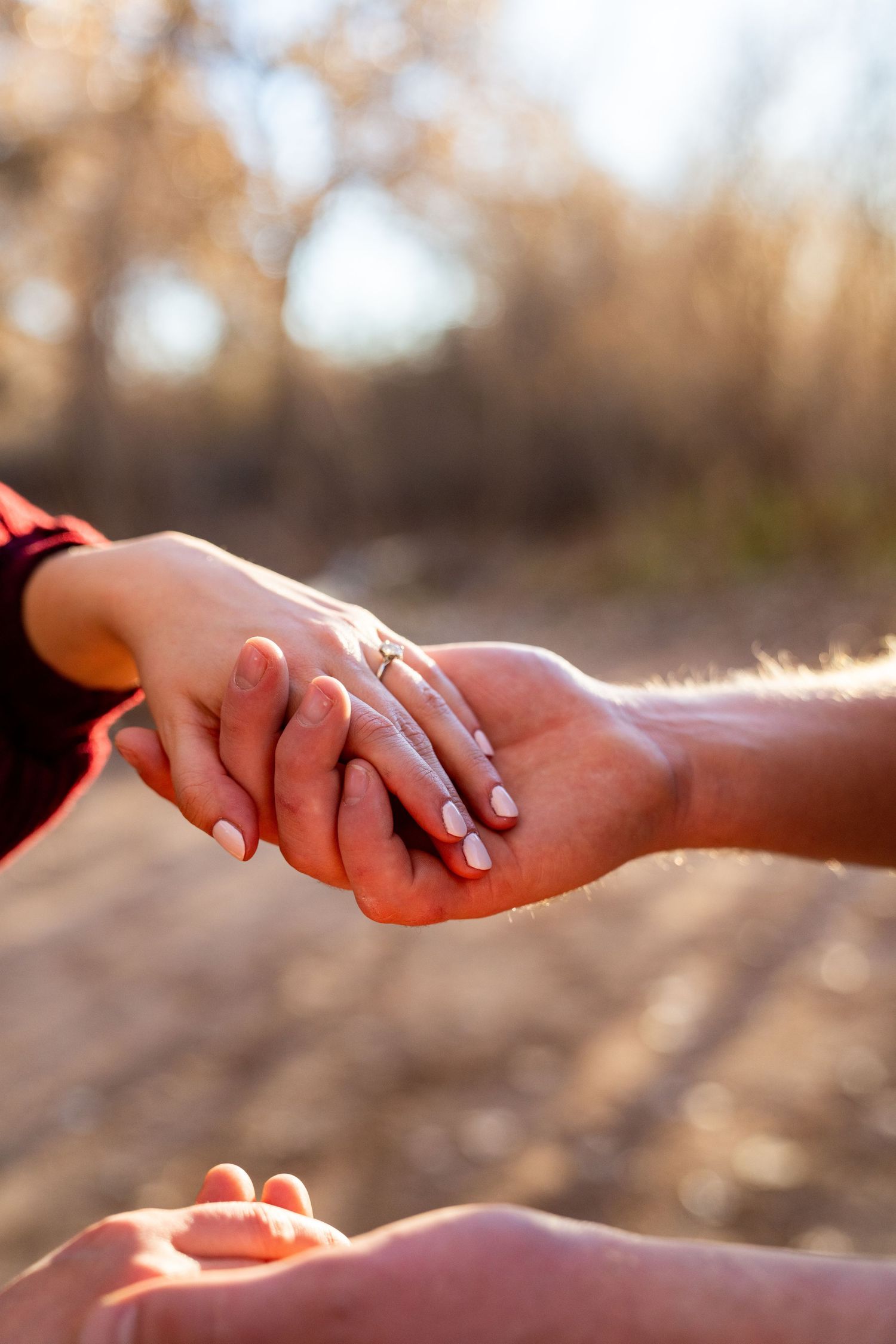 Close-up of couple holding hands outdoors, woman wearing an engagement ring, with warm bokeh background.