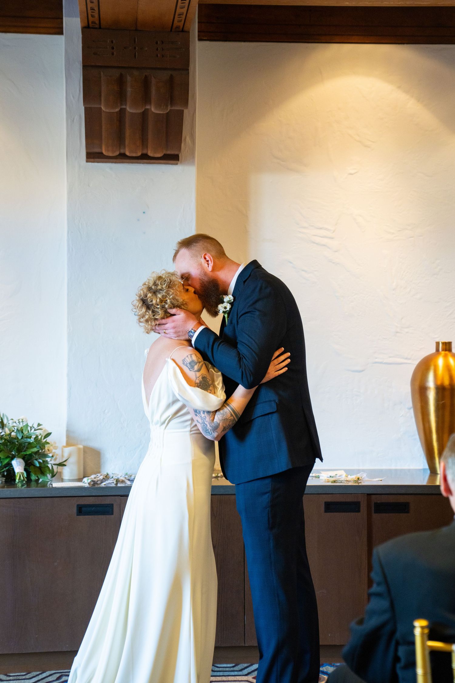 Bride and groom share their first kiss during indoor wedding ceremony, bride in white gown with tattoos, groom in navy suit.