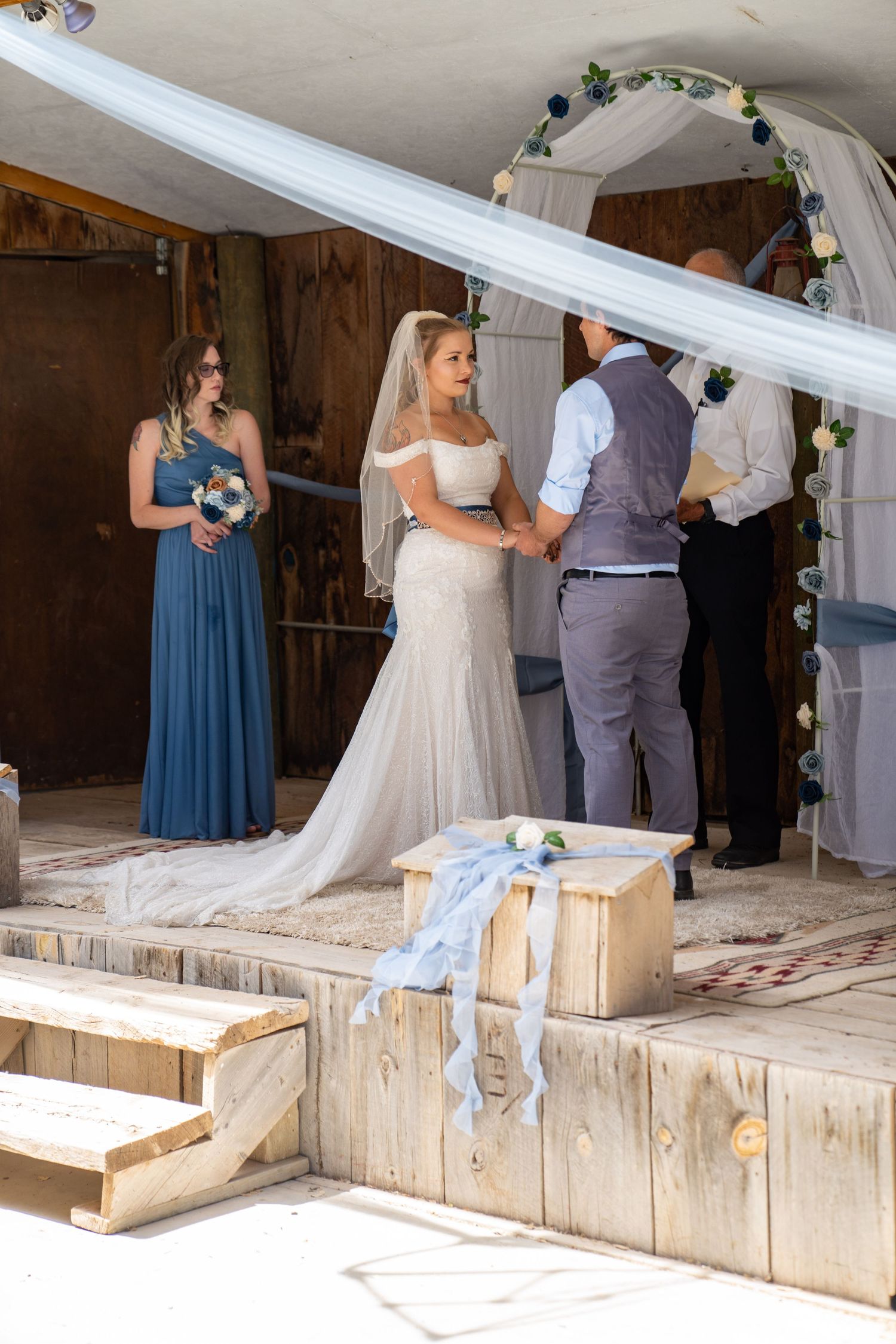 A wedding ceremony takes place on wooden steps under a white draped arch with a bridesmaid in a blue dress.