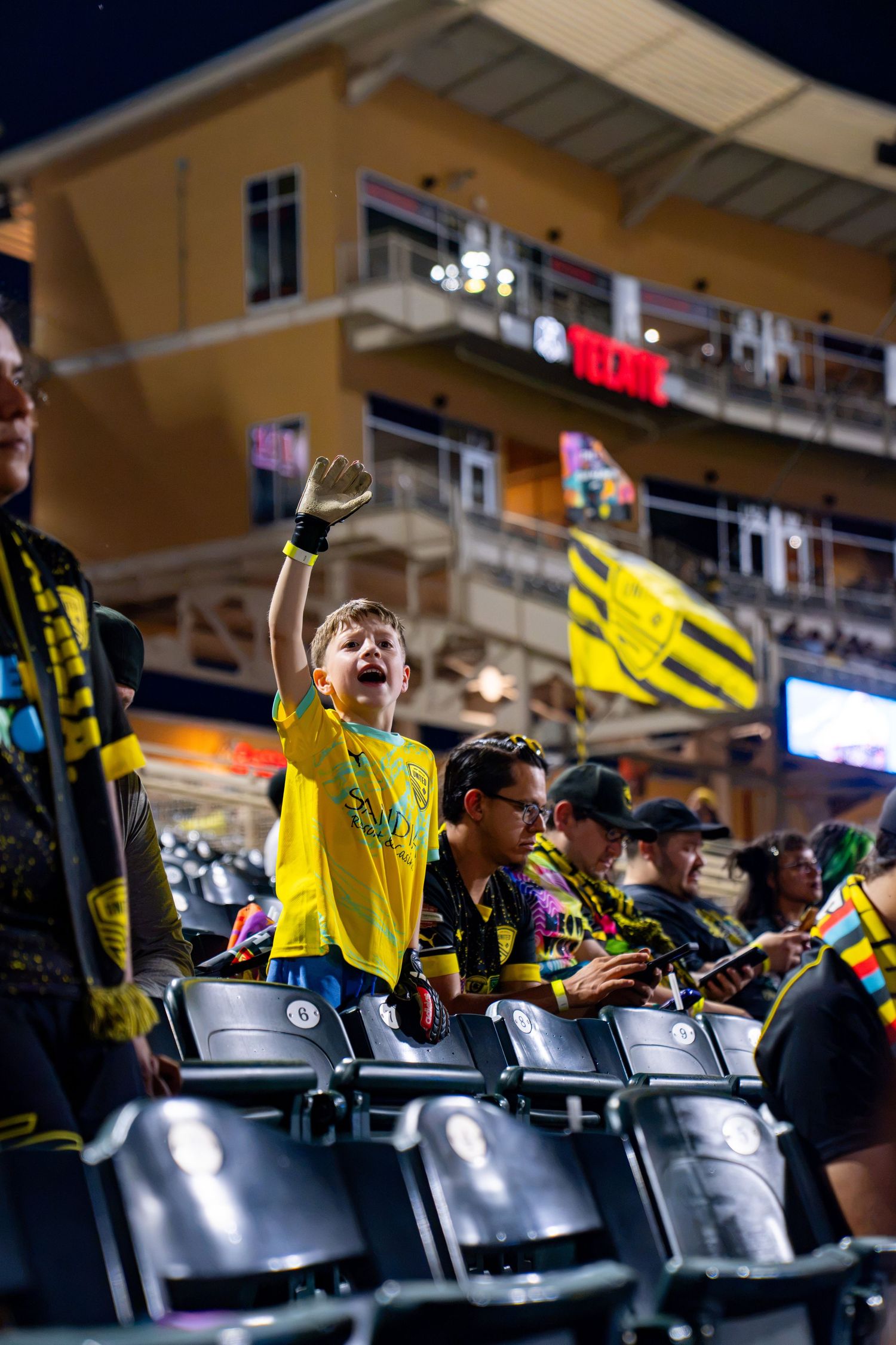 Excited young soccer fan in yellow jersey cheering at an MLS stadium during a night game.