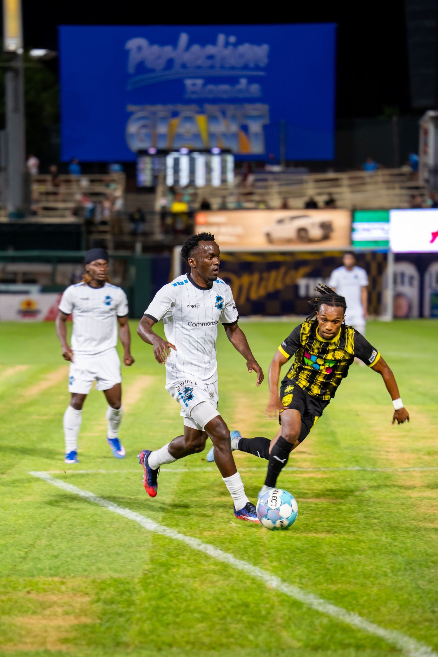 Soccer players compete for the ball during a night match, with one player in white and another in black and yellow jerseys.