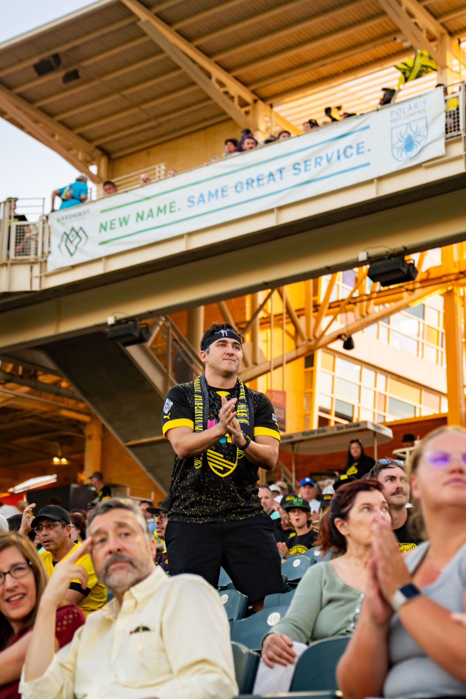Excited fan stands cheering in stadium crowd during a soccer match, with a banner reading New Name Same Great Service.