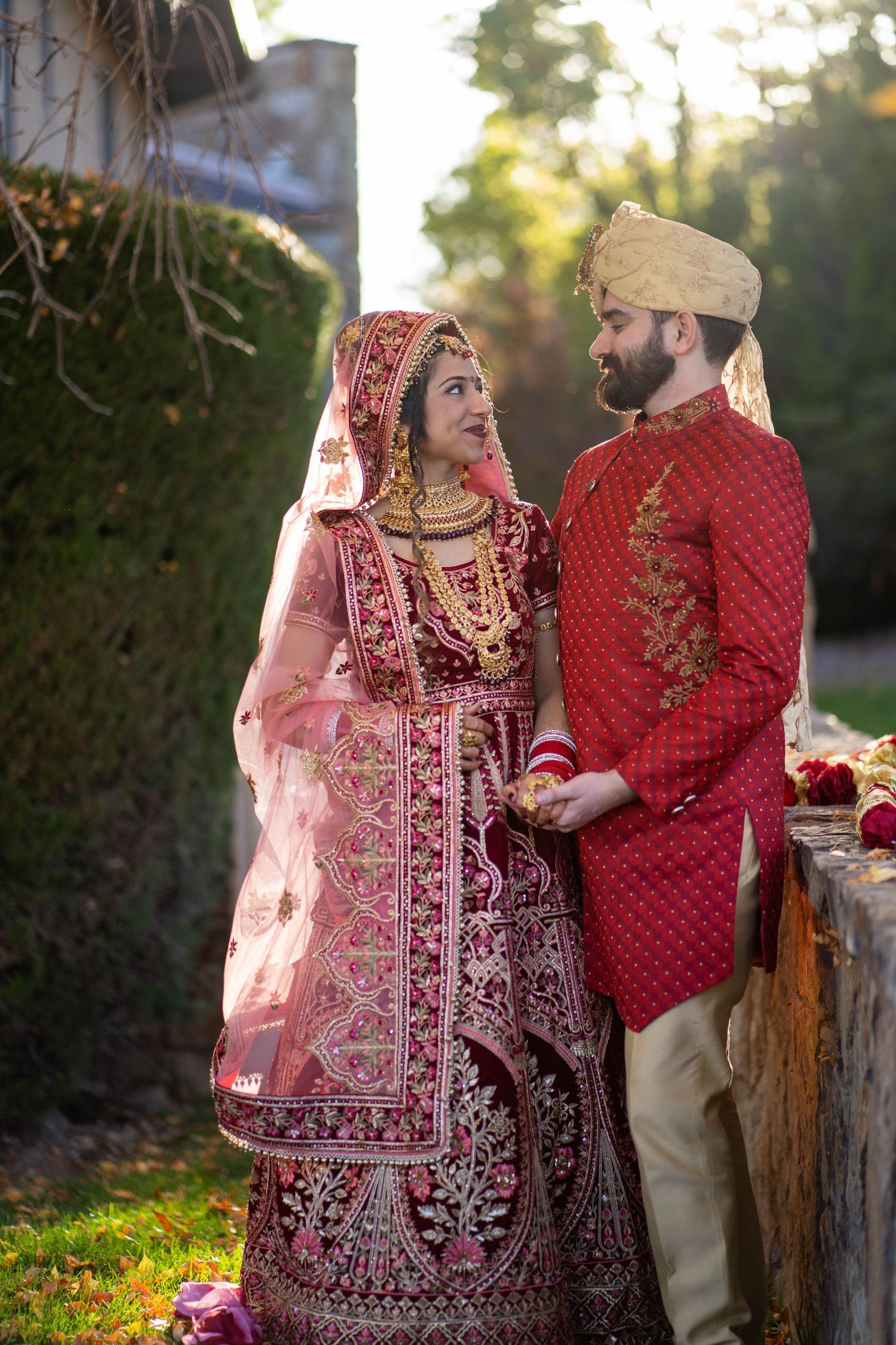 A couple in ornate red and gold Indian wedding outfits stands together in a garden setting.