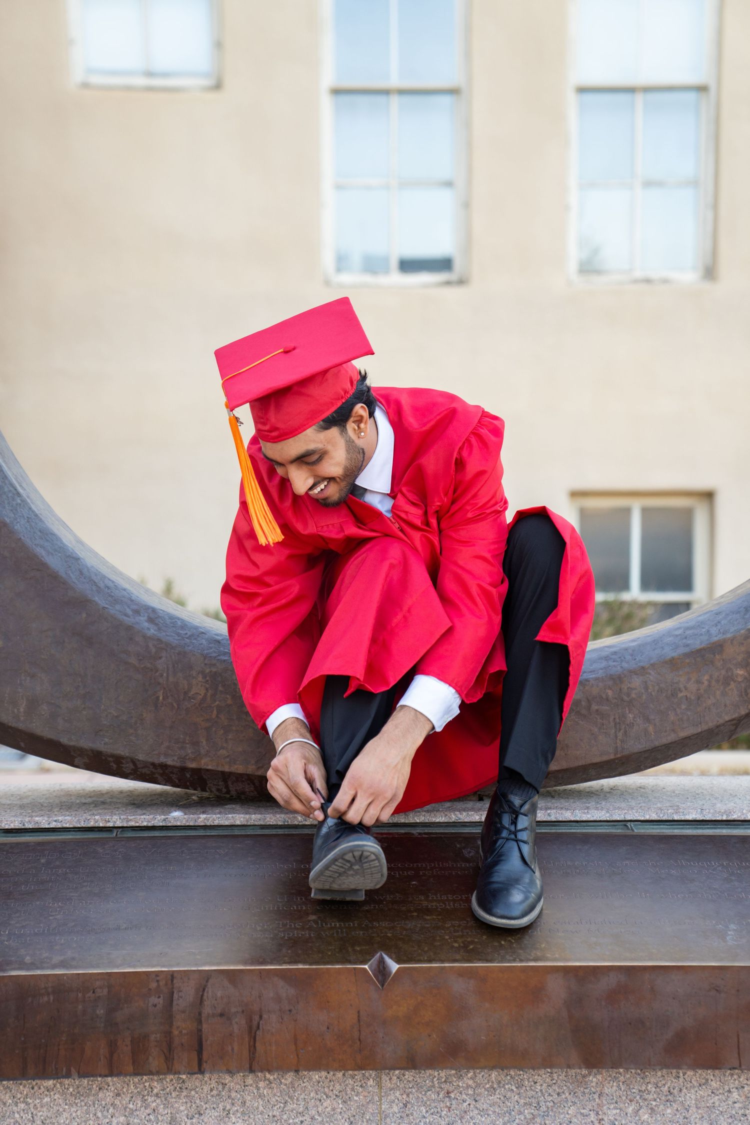 Graduate in bright red cap and gown sits contemplatively on metal structure.