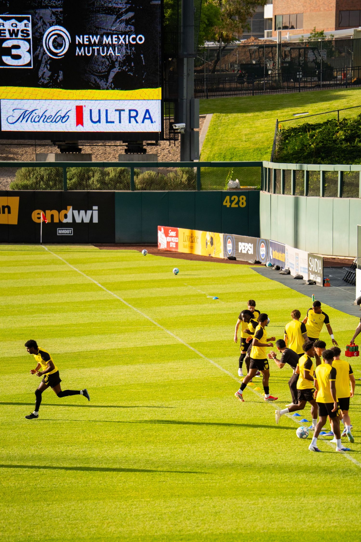 Soccer players in yellow uniforms train on a green field at a stadium with New Mexico Mutual and Michelob Ultra ads.