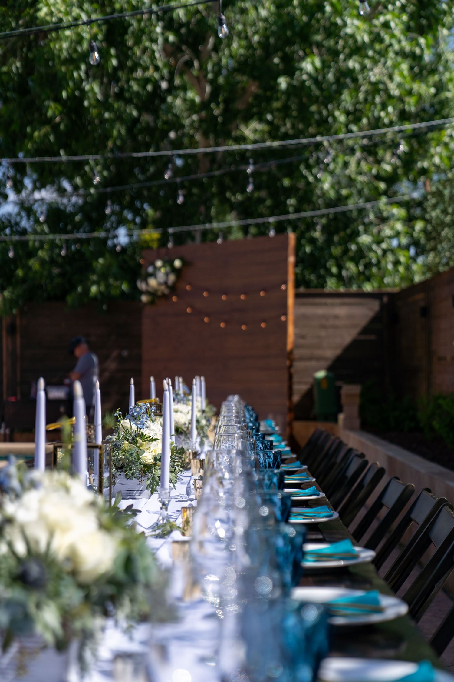 Long outdoor dining table set with elegant place settings featuring turquoise blue napkins and glassware under string lights.