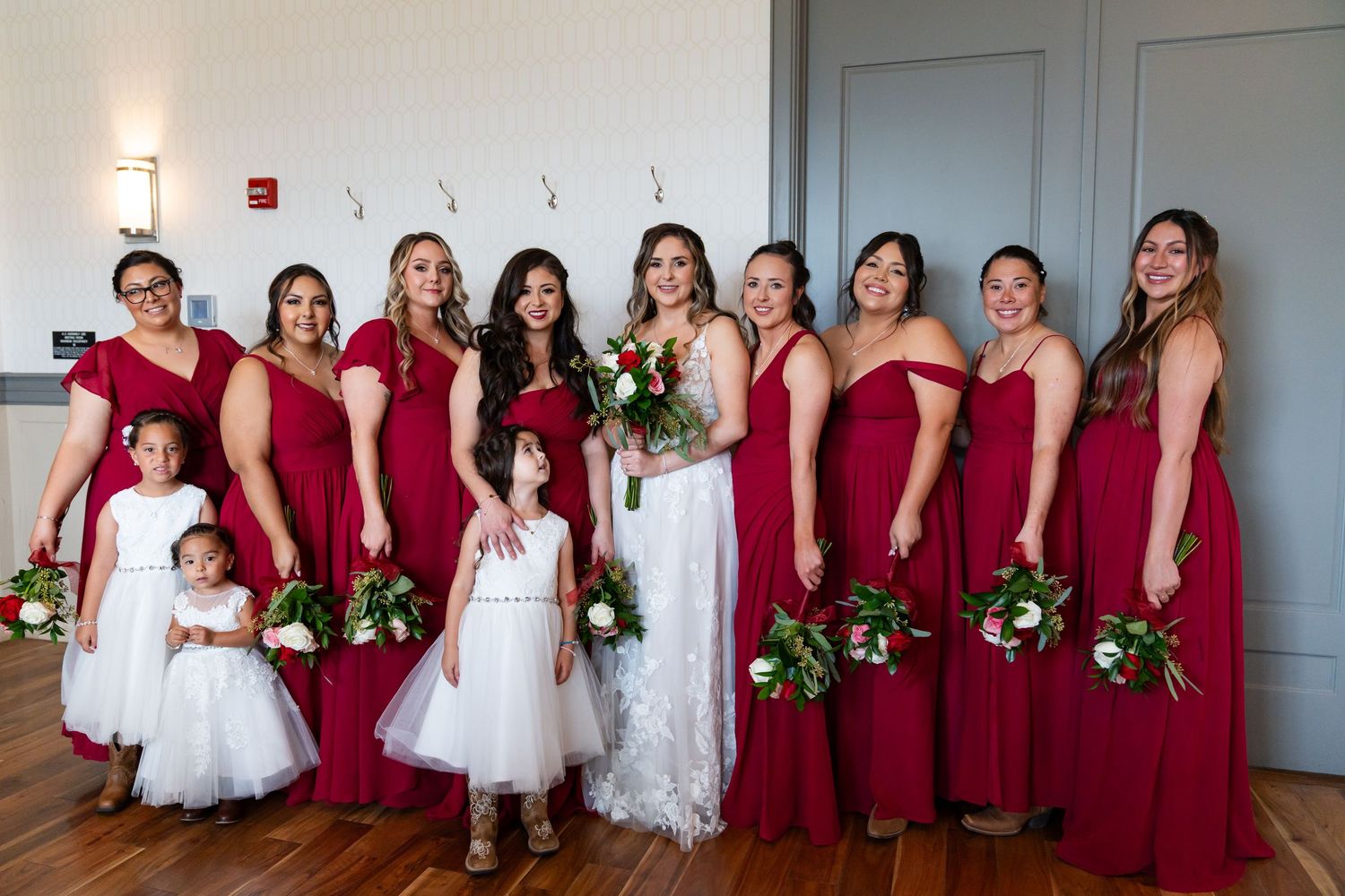 Wedding party poses in red dresses and formal attire against a light gray wall.