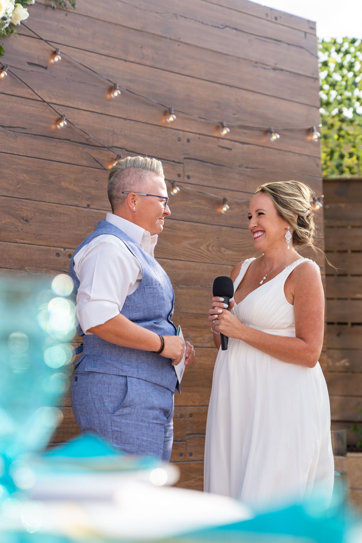 Two people share an emotional moment during a wedding ceremony against a rustic wooden wall with string lights.
