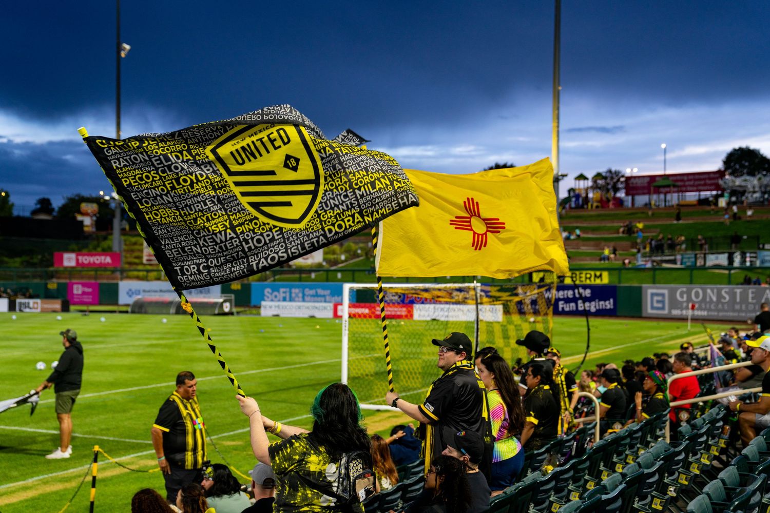 New Mexico United soccer fans wave team and state flags at a vibrant outdoor match under dramatic stormy skies.