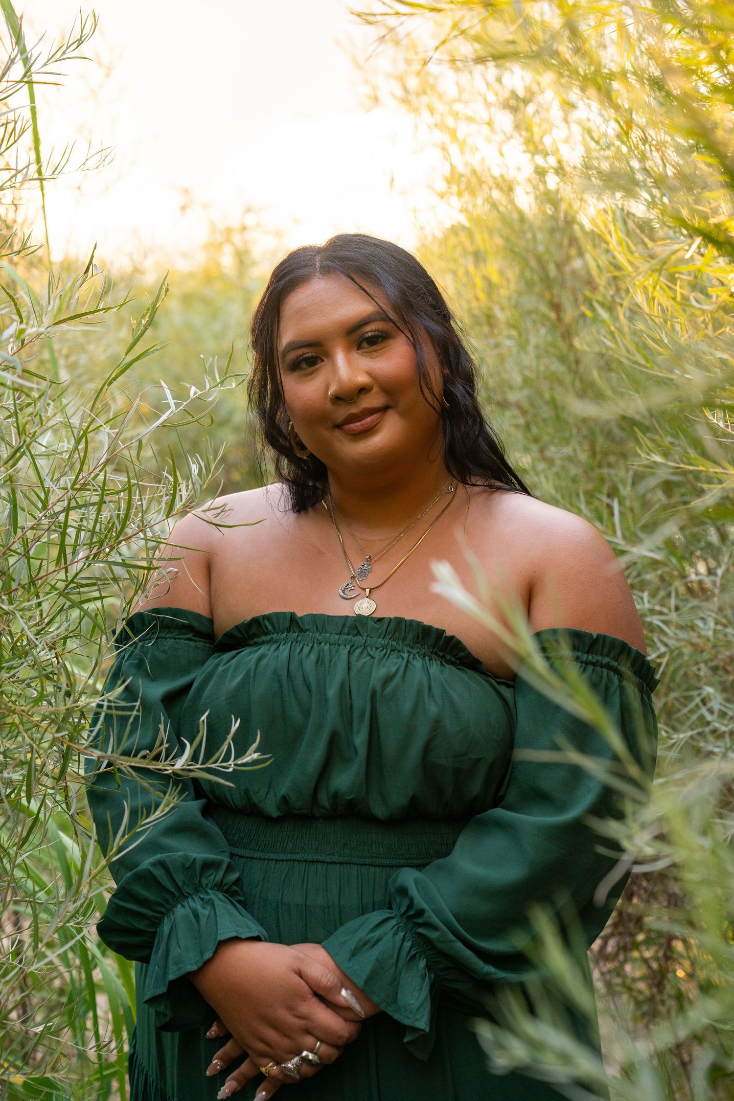Woman in green off-shoulder dress poses among lush greenery in golden hour light, wearing layered necklaces.