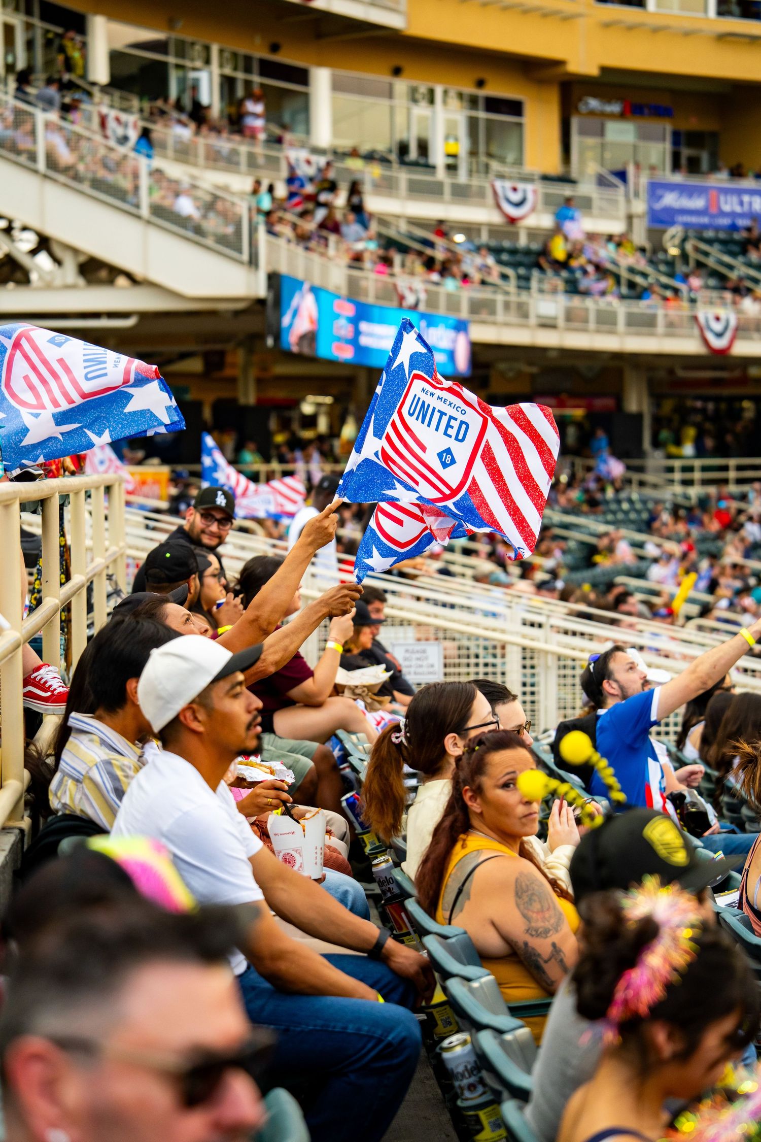 Fans wave New Mexico United flags in stadium stands during a sunny soccer match day.