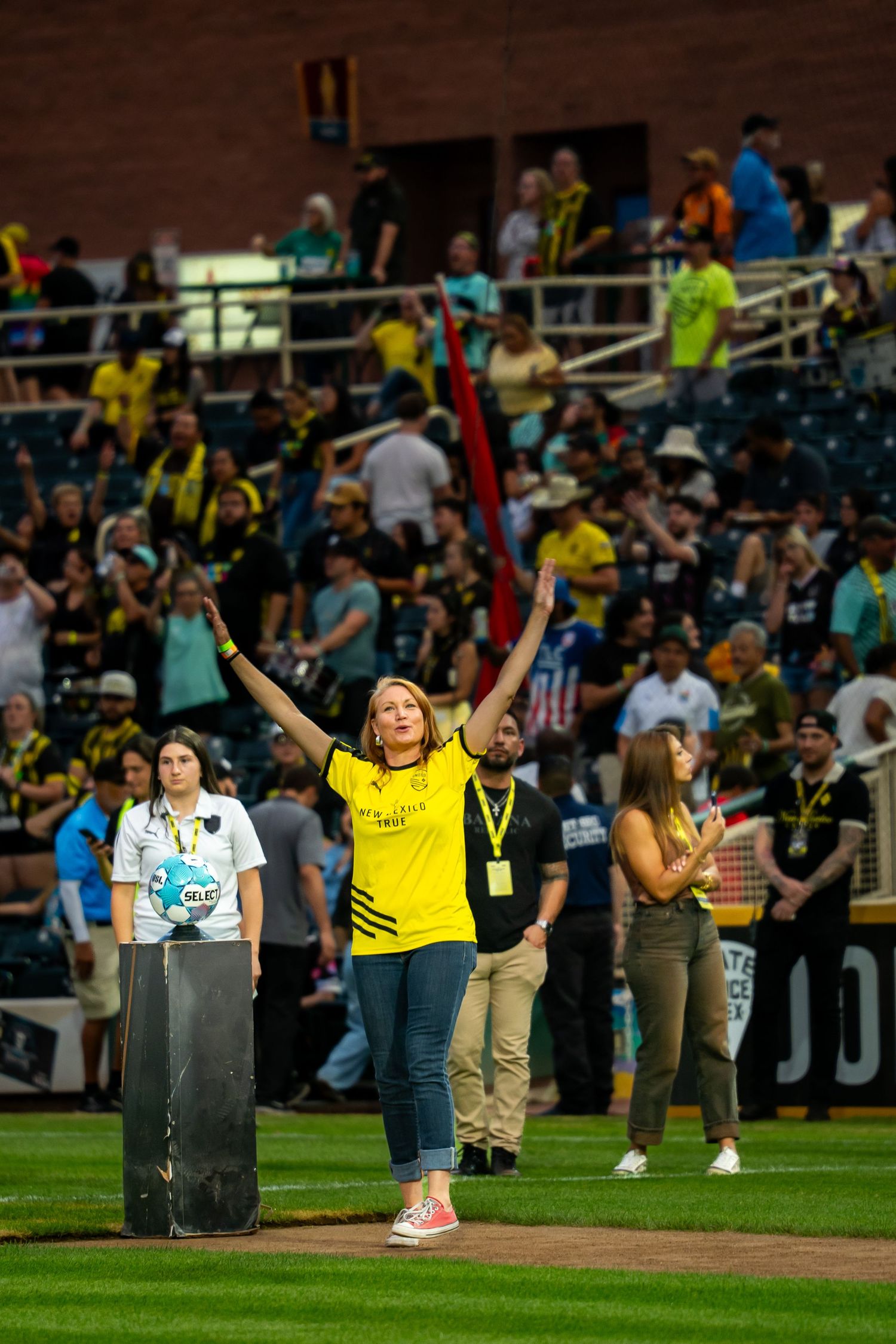 Woman in yellow jersey raises arms at soccer stadium filled with fans in matching yellow team colors.