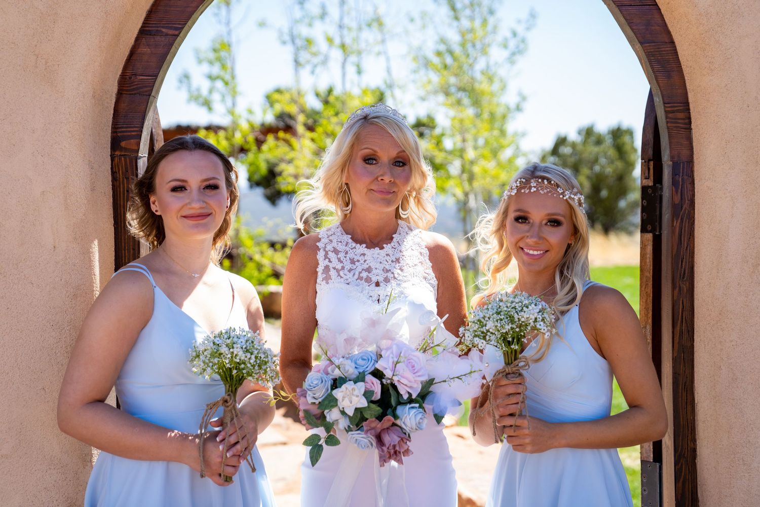 A row of bridesmaids in white dresses holding small bouquets stand together at an outdoor wedding venue.