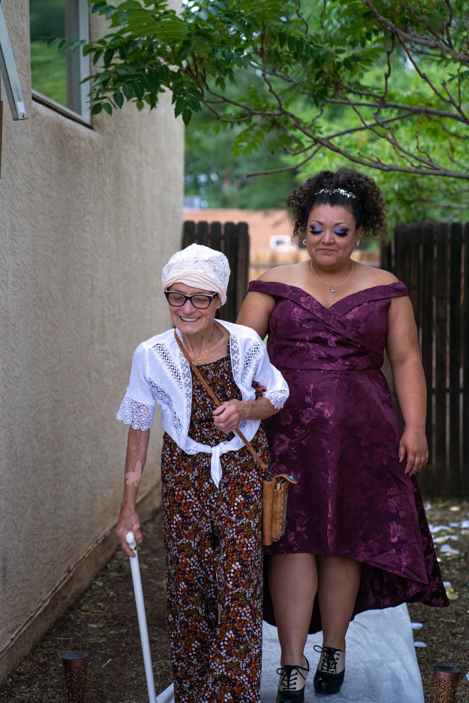 Two people walking together outdoors, one wearing a floral dress and white blouse and another in a purple dress.