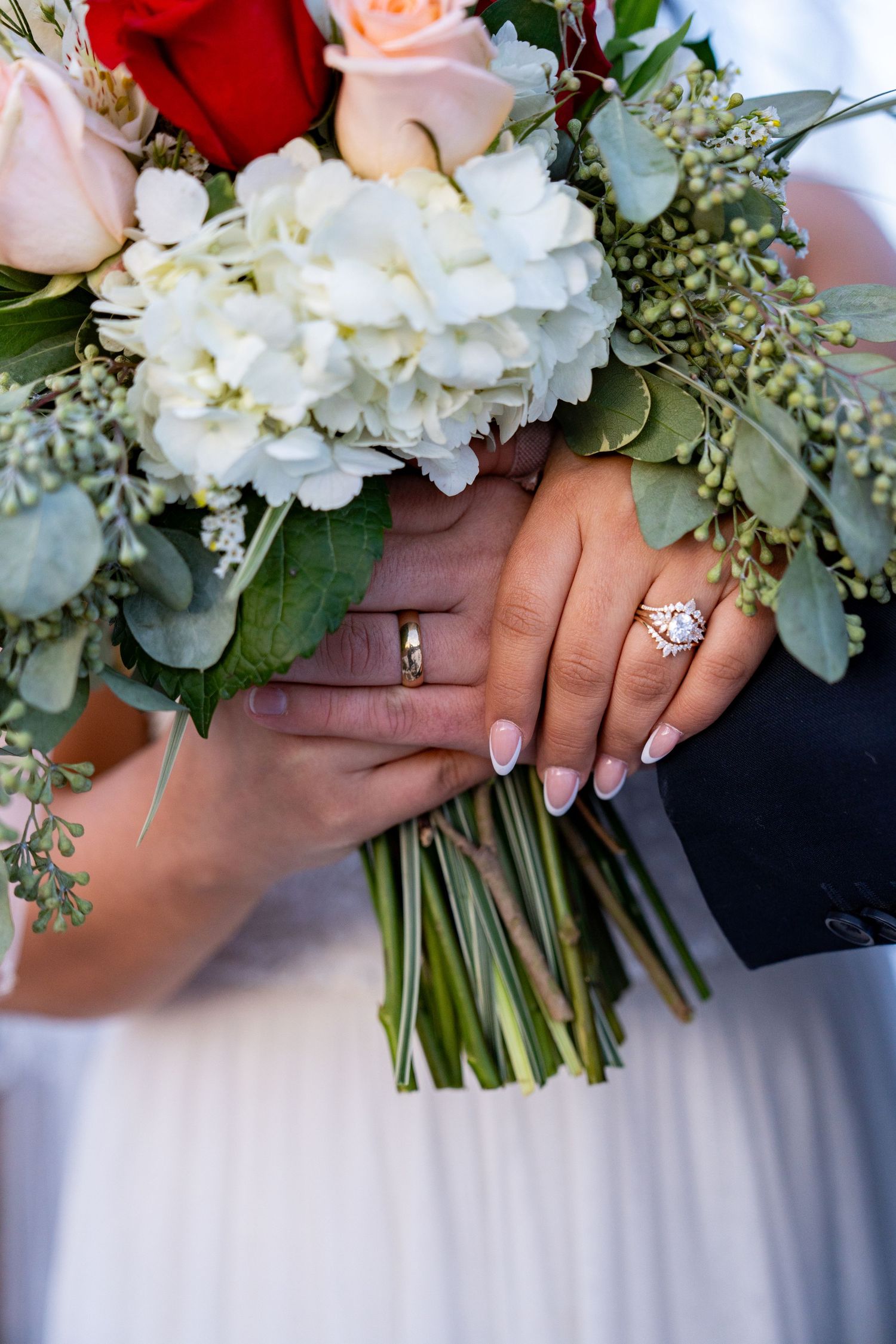 Close-up of wedding rings and hands holding a beautiful bouquet with roses, hydrangeas, and greenery.