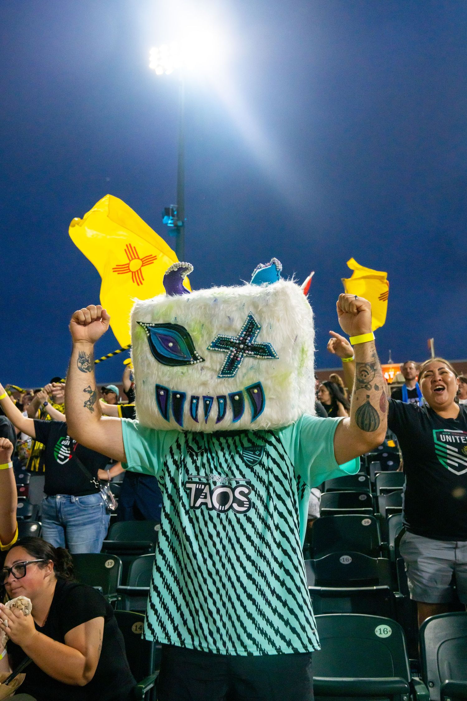 New Mexico United fan wears large furry monster mascot head with Taos jersey at night game.
