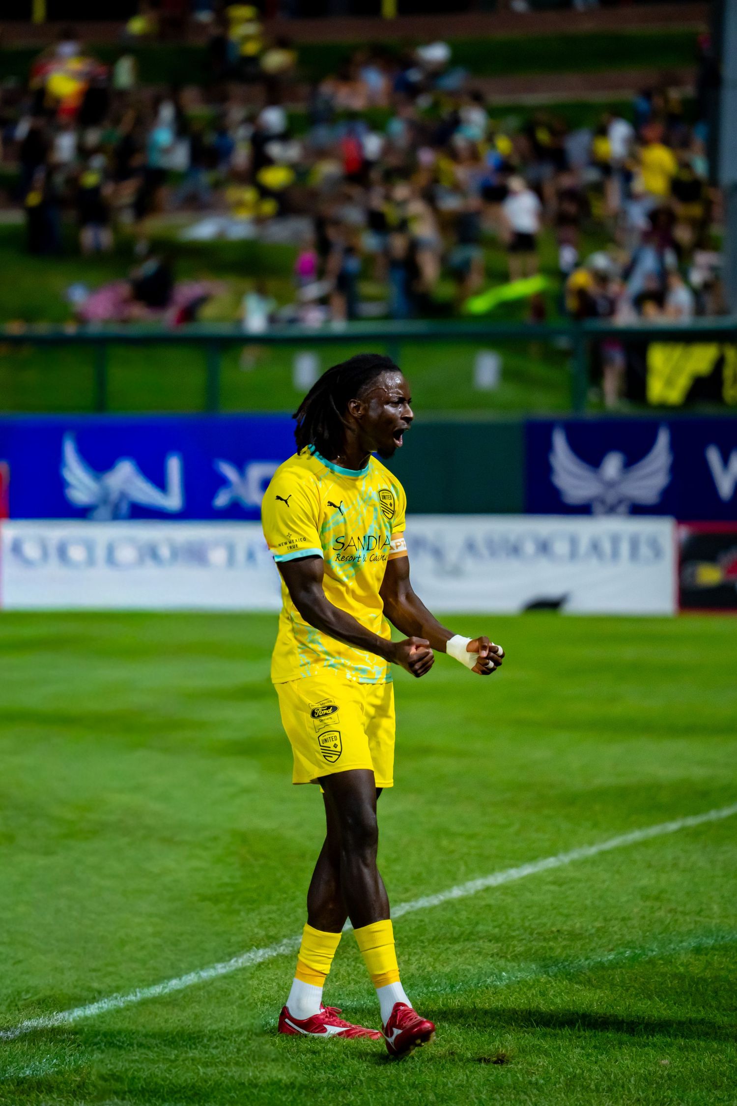 Soccer player in yellow kit celebrates on field during night match with crowd in background.