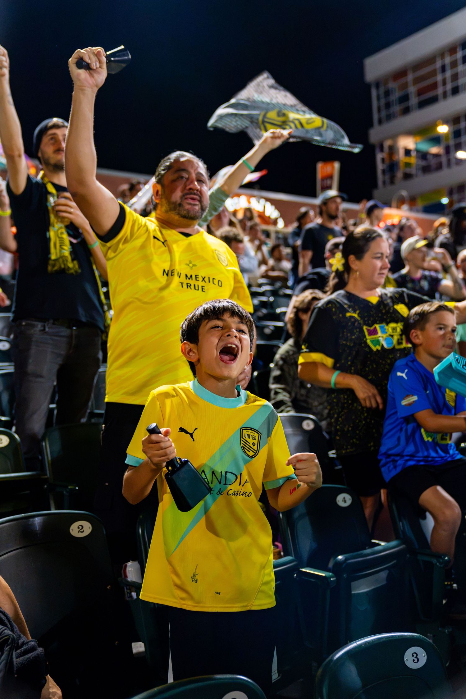 Excited New Mexico United soccer fans cheering in yellow jerseys at a night game, including an enthusiastic young boy.