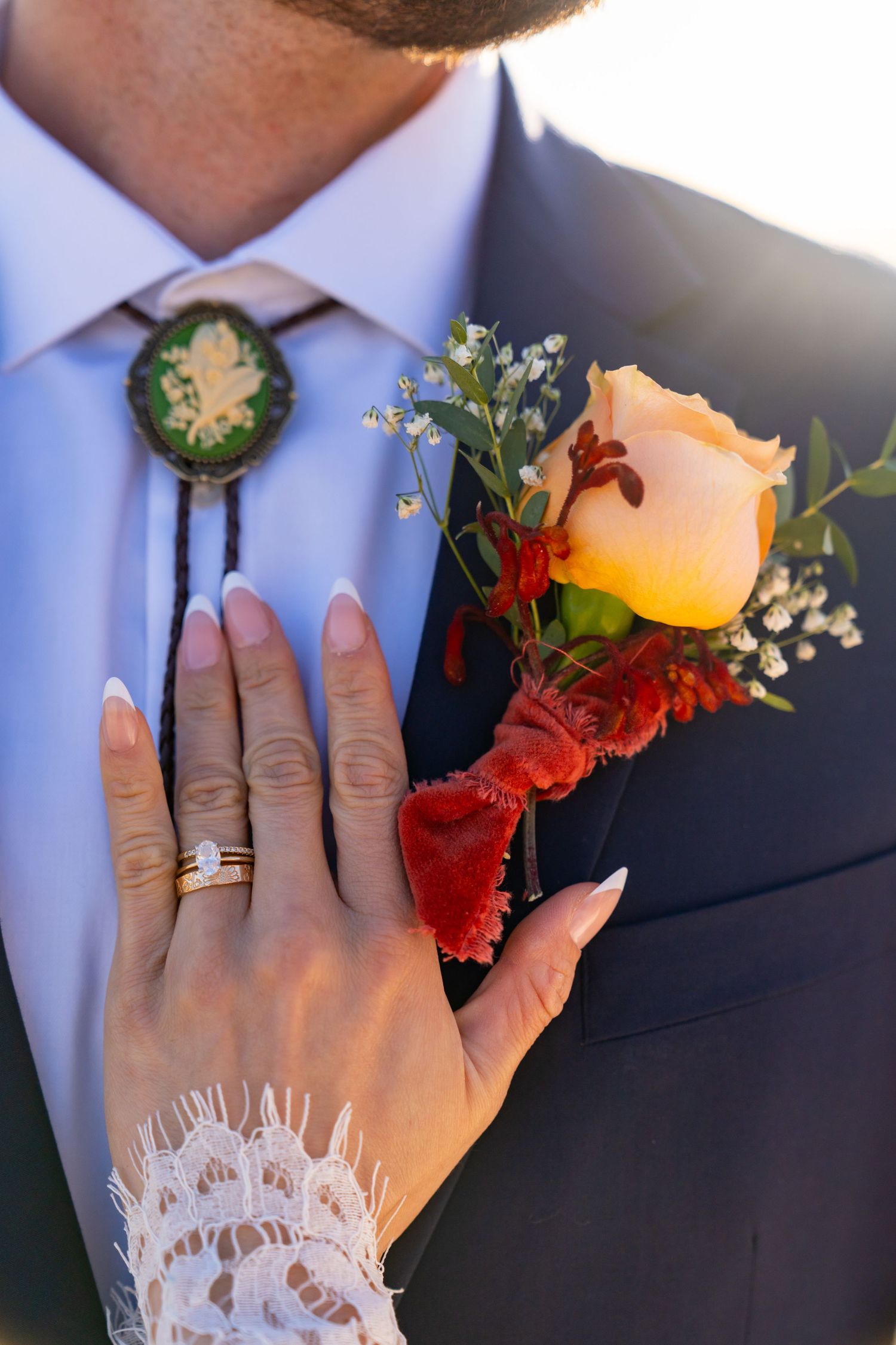 Close-up of bride's hand with wedding ring touching groom's boutonniere with peach rose and bolo tie.