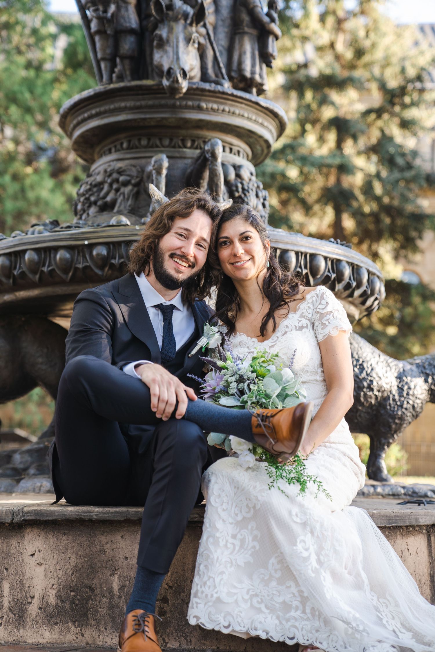 A newlywed couple sits on the edge of an ornate stone fountain in a garden setting.