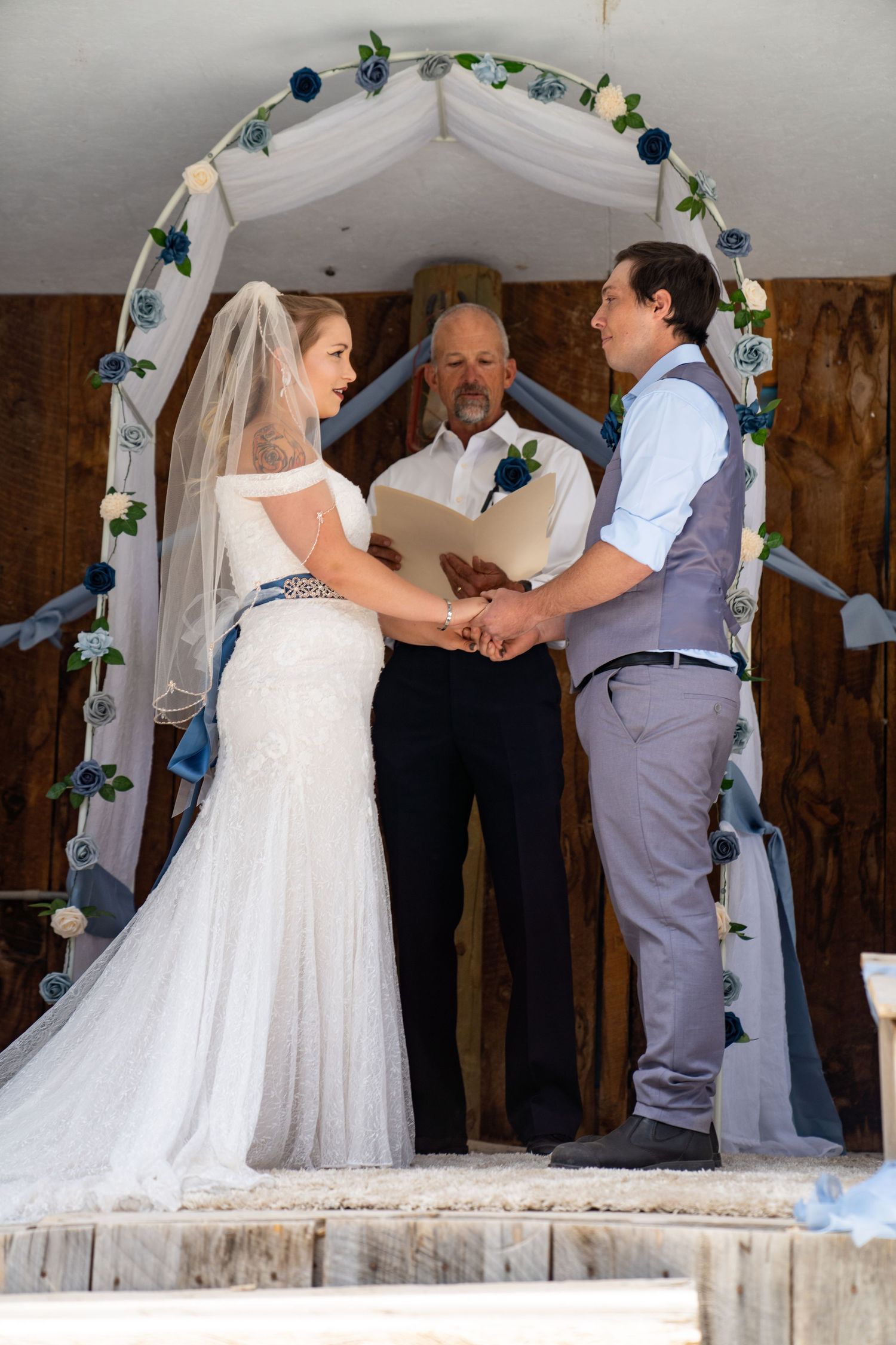 A wedding ceremony takes place under a decorated arch with blue and white floral accents in a rustic barn setting.