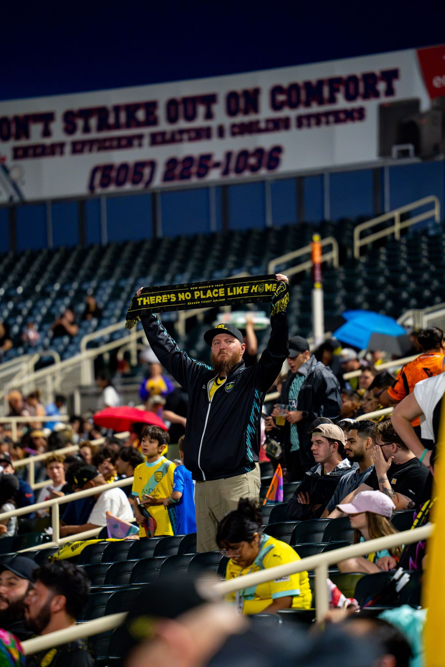Fan holds scarf reading There's No Place Like Home at a crowded outdoor sports stadium.