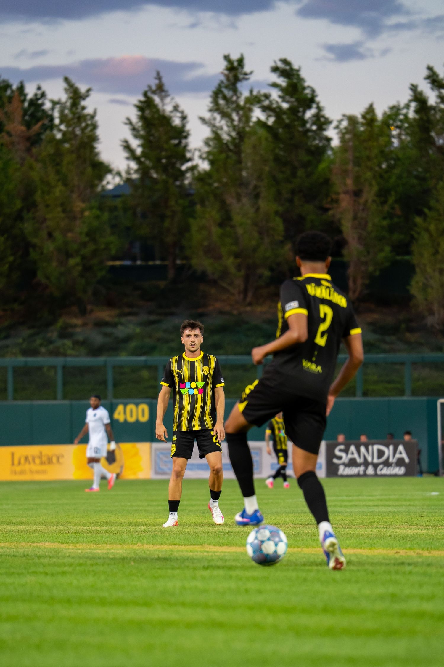 Soccer players in yellow and black uniforms compete on a green field near Sandia Resort, with trees in the background.