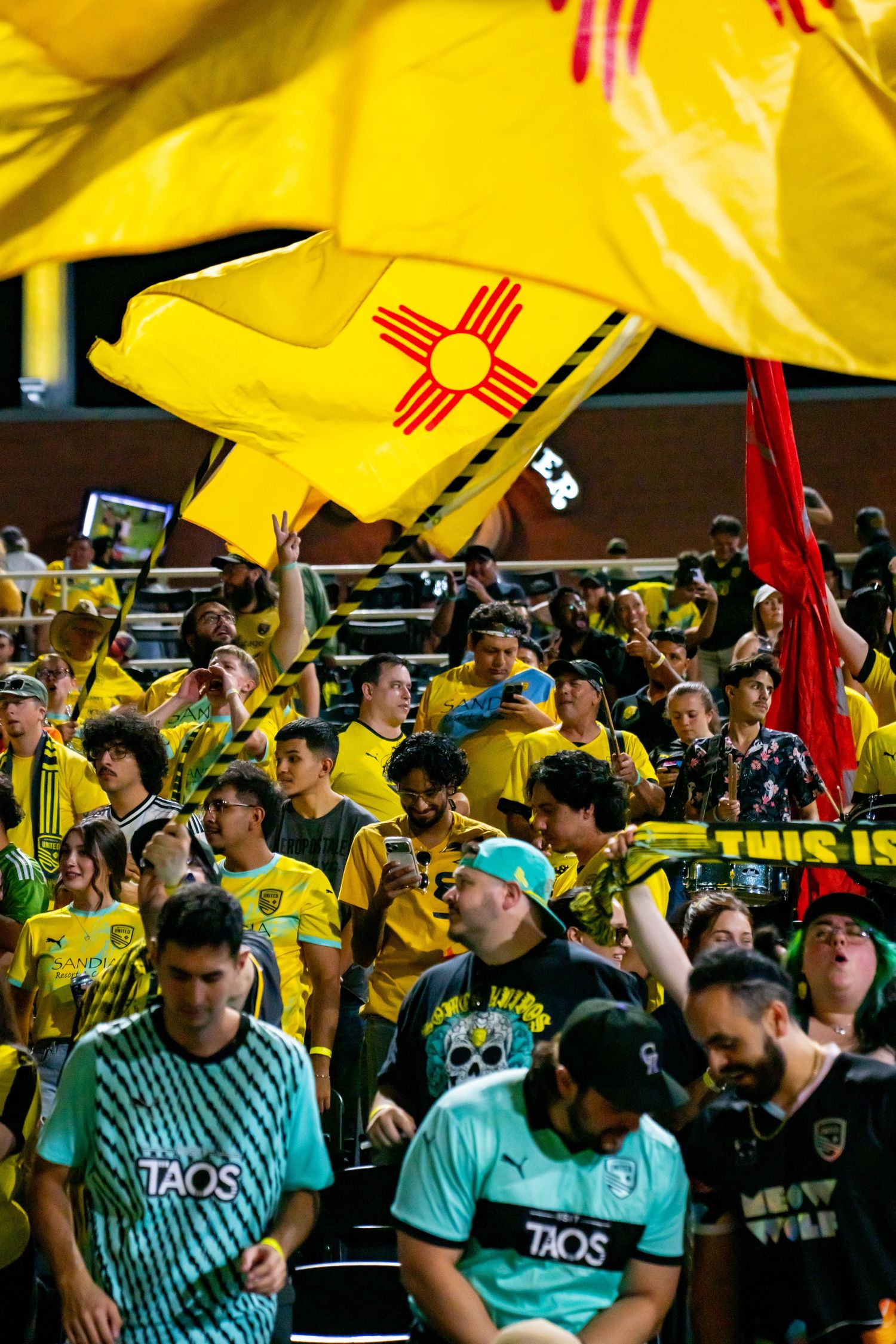 New Mexico United soccer fans wave the state flag in yellow and black gear at a lively stadium match.