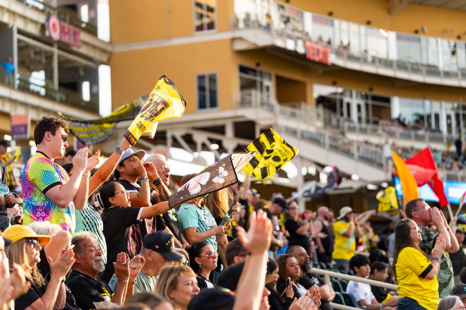 Excited soccer fans wave yellow and black flags and noisemakers in packed stadium stands during a lively match.